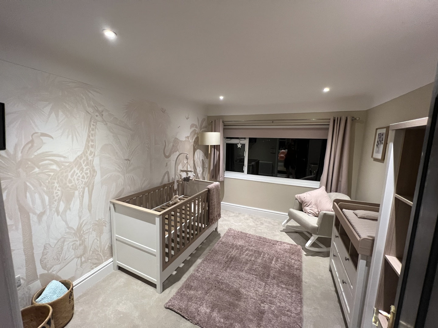 A cozy children’s room features a crib, changing table, armchair with pillow, soft rug, and the Magic Jungle Wallpaper Mural with giraffes and trees. Soft lighting by the large window and curtains creates a warm, inviting atmosphere.