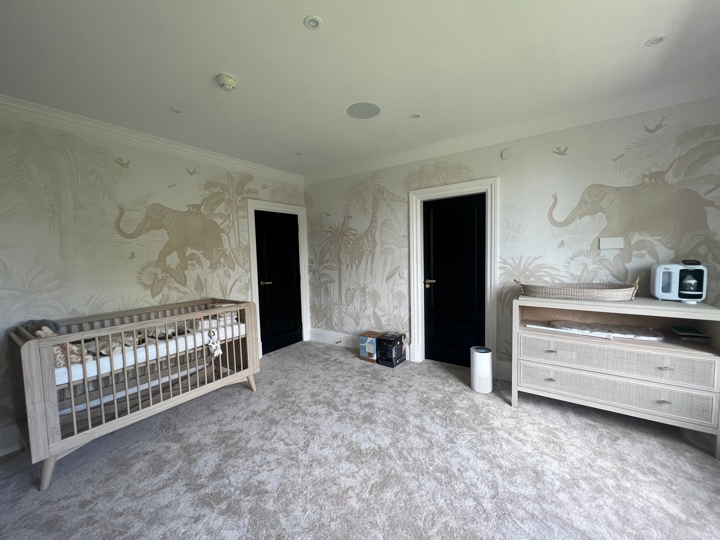 A spacious kids room with light carpet, a crib on the left, a changing table and baby bottle maker on the right, two black doors, and a Magic Jungle Wallpaper Mural with playful animals and trees.