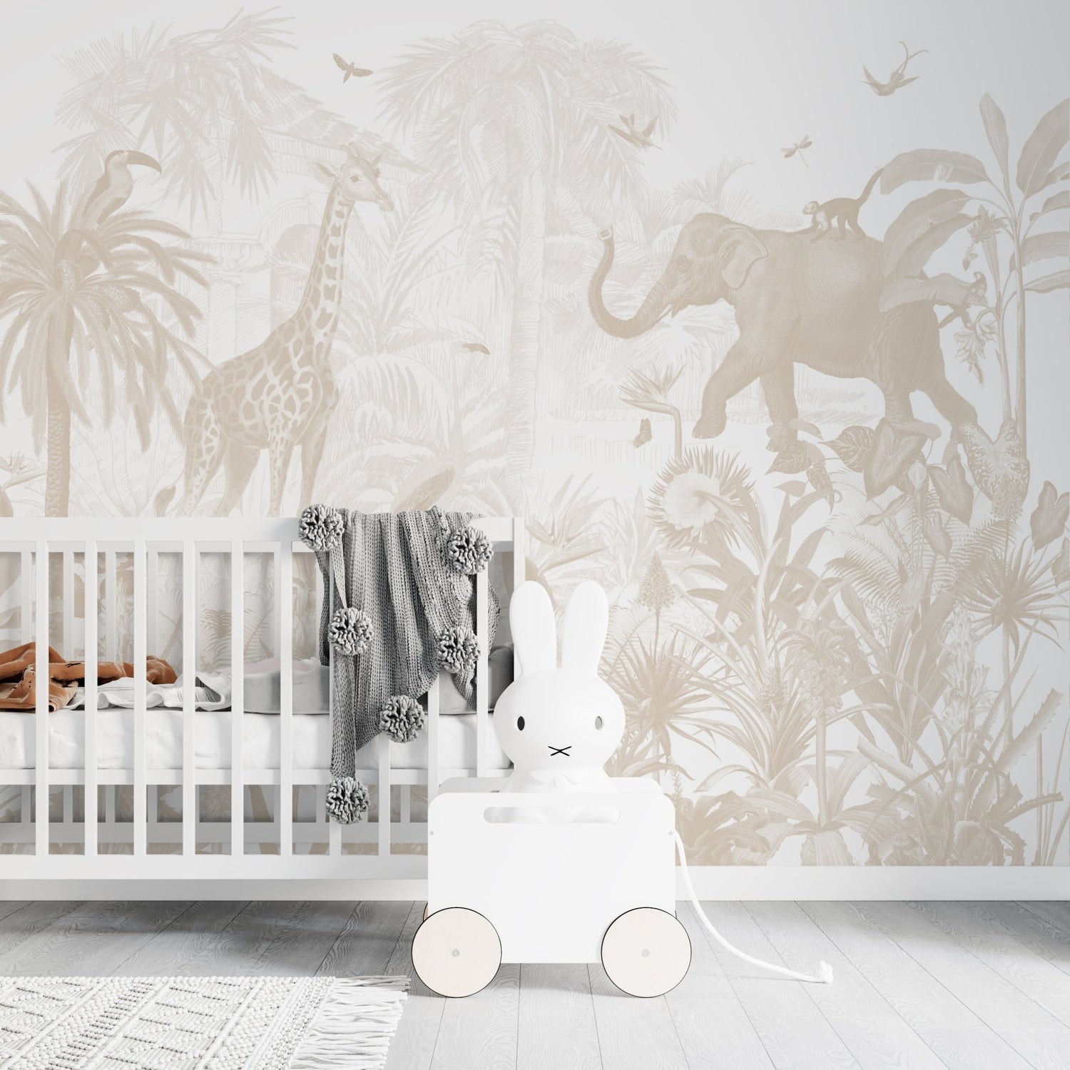 A nursery with a white crib, gray blanket, and white bunny toy on wheels. The Magic Jungle Wallpaper Mural features giraffes, elephants, monkeys, and birds in neutral tones—ideal for a charming childrens room.