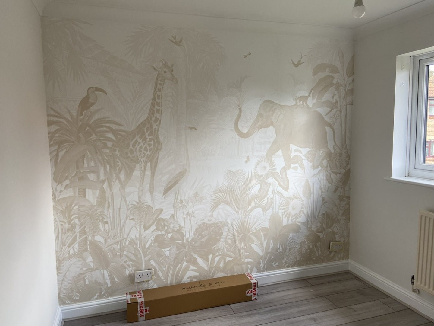 A small kids’ room with light wood floors features the Magic Jungle Wallpaper Mural, showcasing a giraffe, elephant, and tropical plants in neutral tones. A long cardboard box sits against the wall beneath the mural.