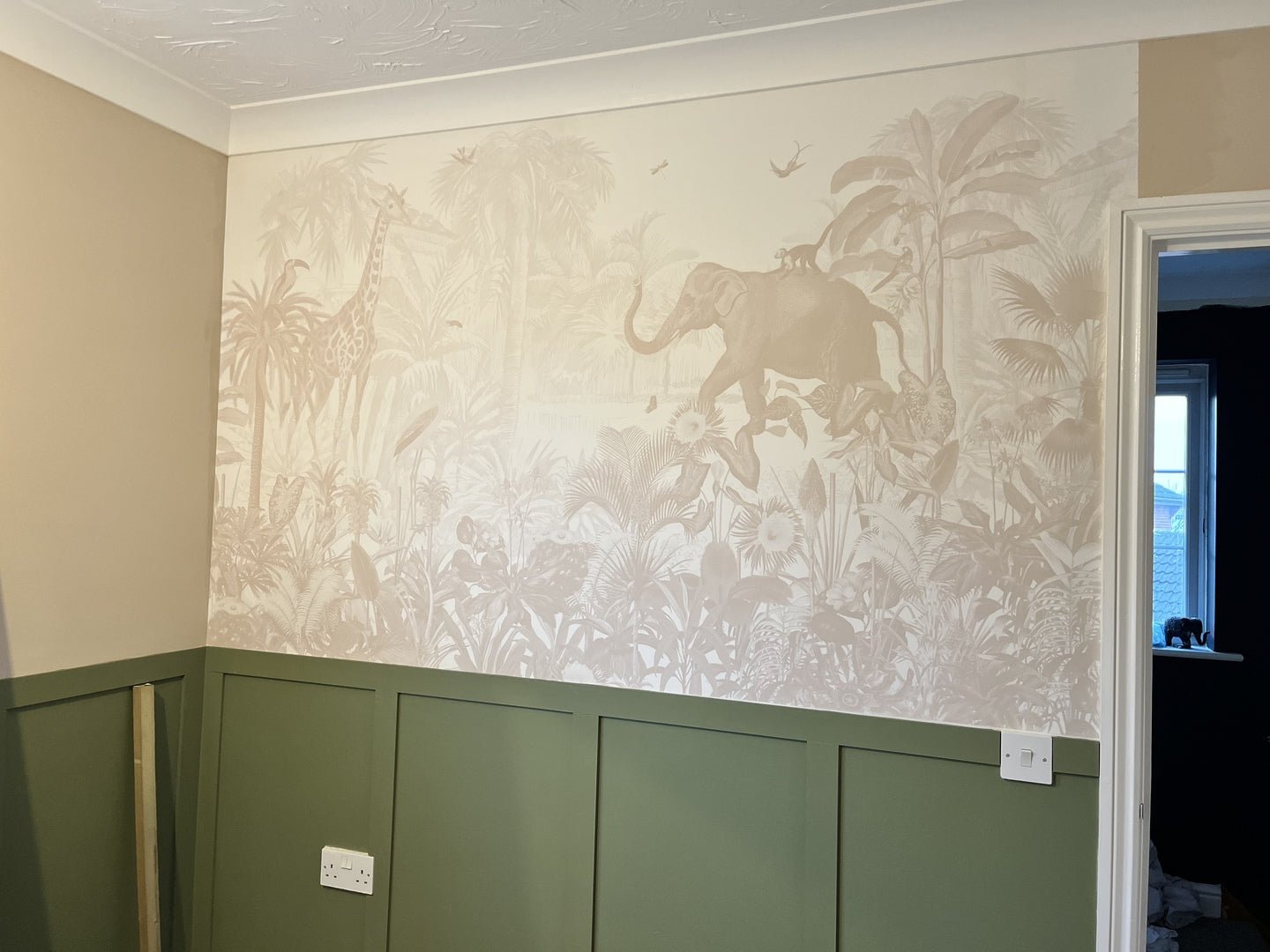 A children’s room features the Magic Jungle Wallpaper Mural—beige with animals like an elephant and giraffe, tropical plants, and birds—above green paneling, against neutral walls with a doorway leading to a darker room.