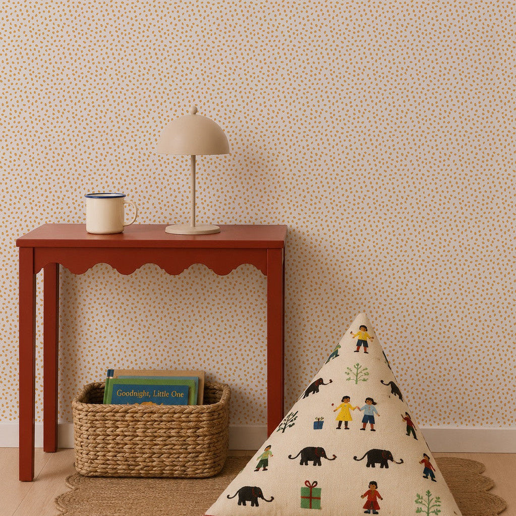 A red table holds a lamp and mug next to a woven basket with books. A triangular cushion featuring colorful people and animal designs sits on a round rug against a statement wall adorned with the Sprinkle Wallpaper in Mustard.