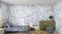 A cozy bedroom features a single bed with blue bedding, Nelly Leopard Print wallpaper sample by munks and me in blue and white. A green cabinet with plants, decor, large oval mirror, garland, pouf, and textured rug adds charm. Natural light enhances the rooms colors.