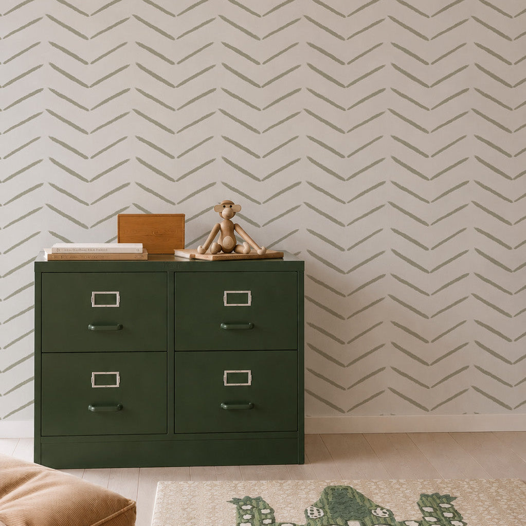 A green drawer cabinet sits against olive herringbone wallpaper, with a toy monkey and boxes on top. A crocodile-patterned rug and tan cushion rest on a light wood floor, creating a space enhanced by stylish wall decor.