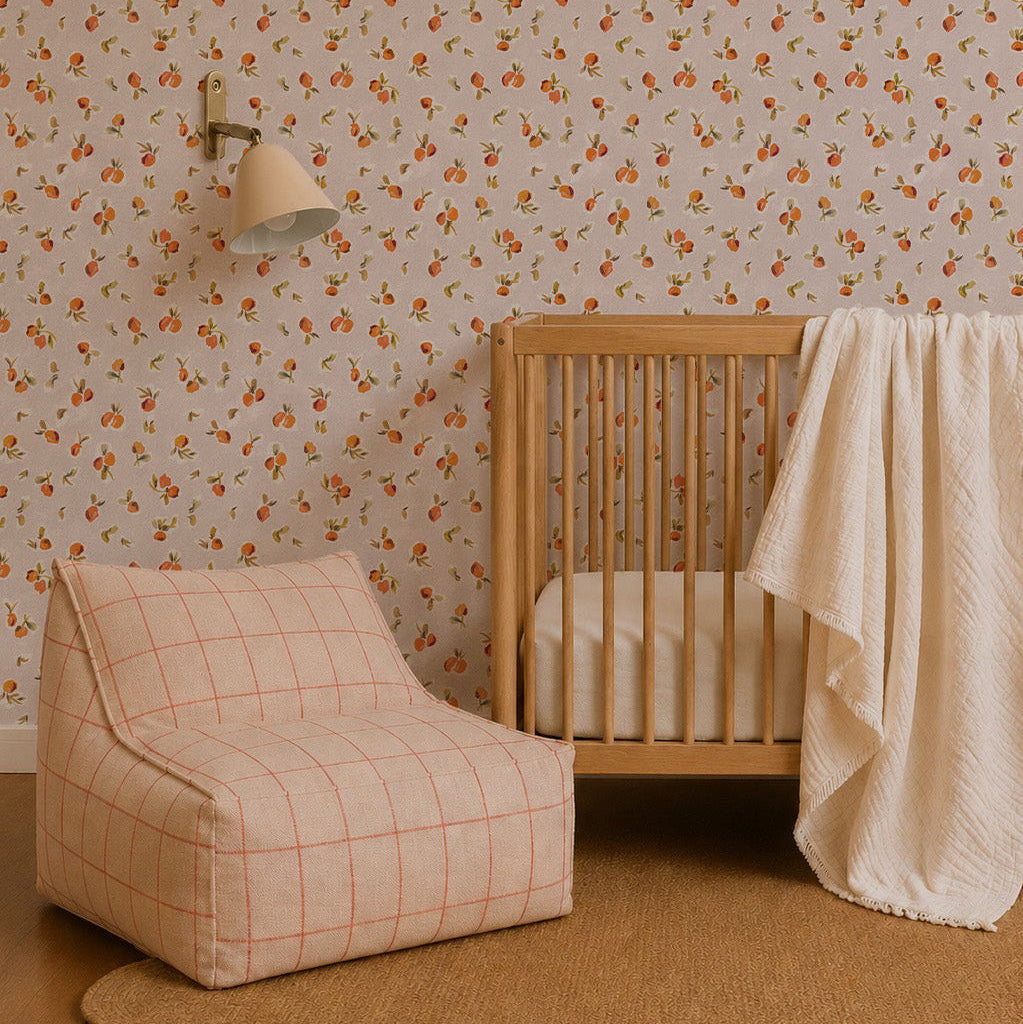 A cozy nursery with Ollies Oranges Wallpaper in pink, a wooden crib draped with a cream blanket, a modern beige and orange plaid lounge chair, a round woven rug, and a wall-mounted lamp.