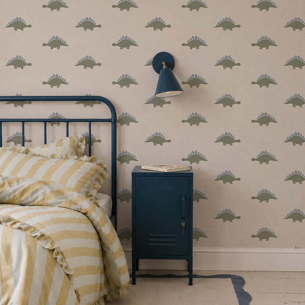 A cozy bedroom features a blue metal bed with yellow-striped bedding, a blue nightstand, a blue wall lamp, and Otis Dinosaur Wallpaper—perfect for playful home decor.