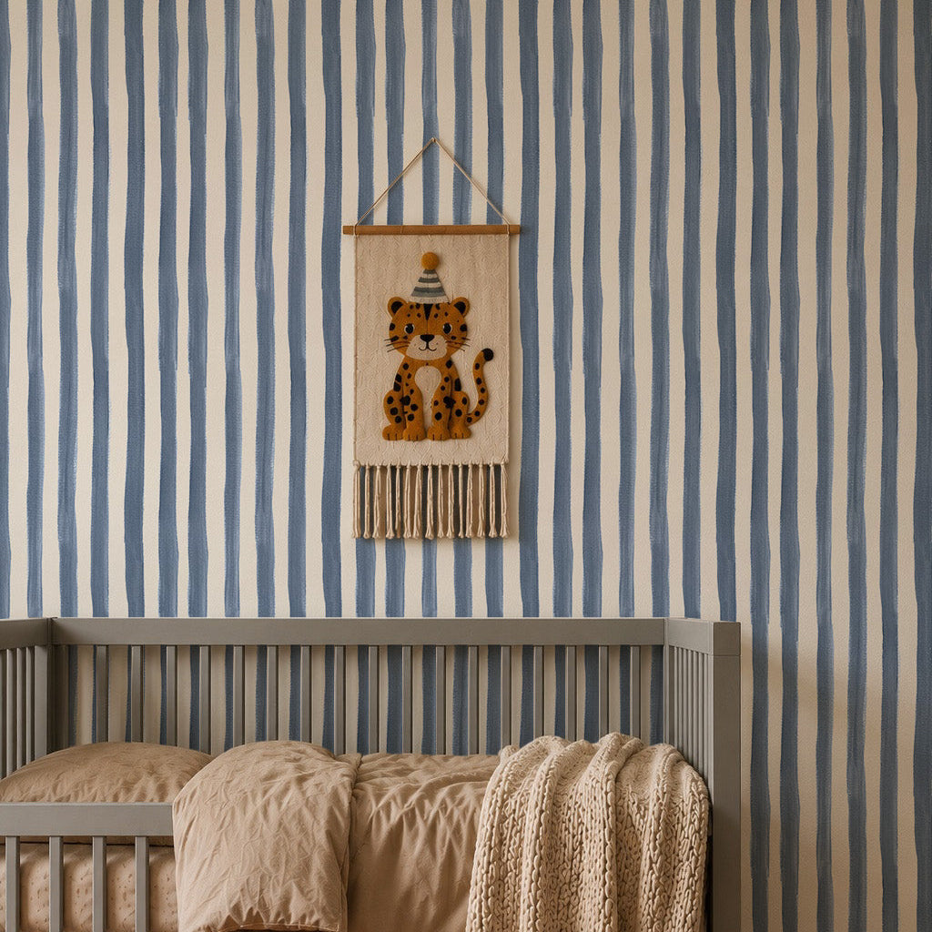 A cozy nursery with a gray crib, tan bedding, knitted beige blanket, rust-colored rug, and wooden toys. The wall features Painted Stripe Wallpaper in Blue and a hanging leopard decoration inspired by Stephanie Breeze.