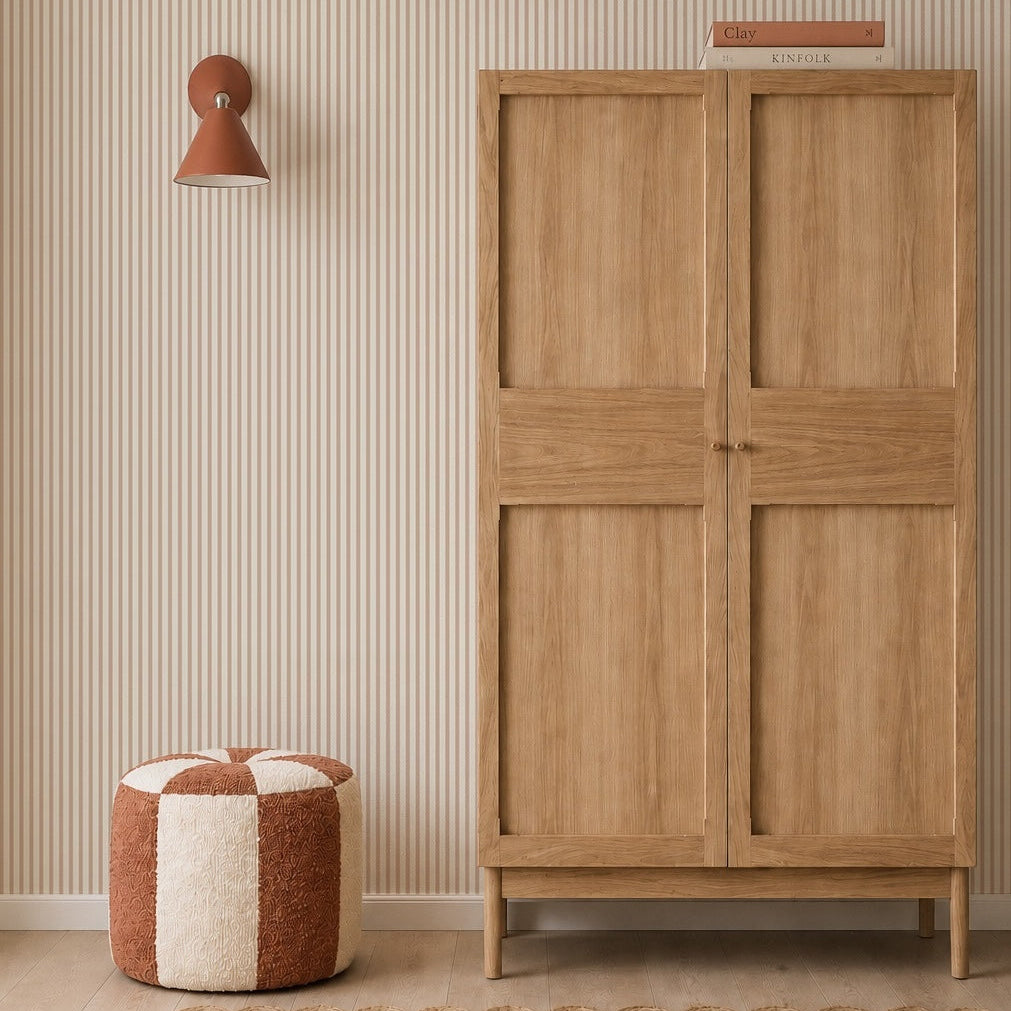 A minimalist room with a wooden wardrobe, a striped cream and rust pouf, woven rug, rust wall sconce, and Pico Stripe Wallpaper in Rose. Two books are stacked on the wardrobe.