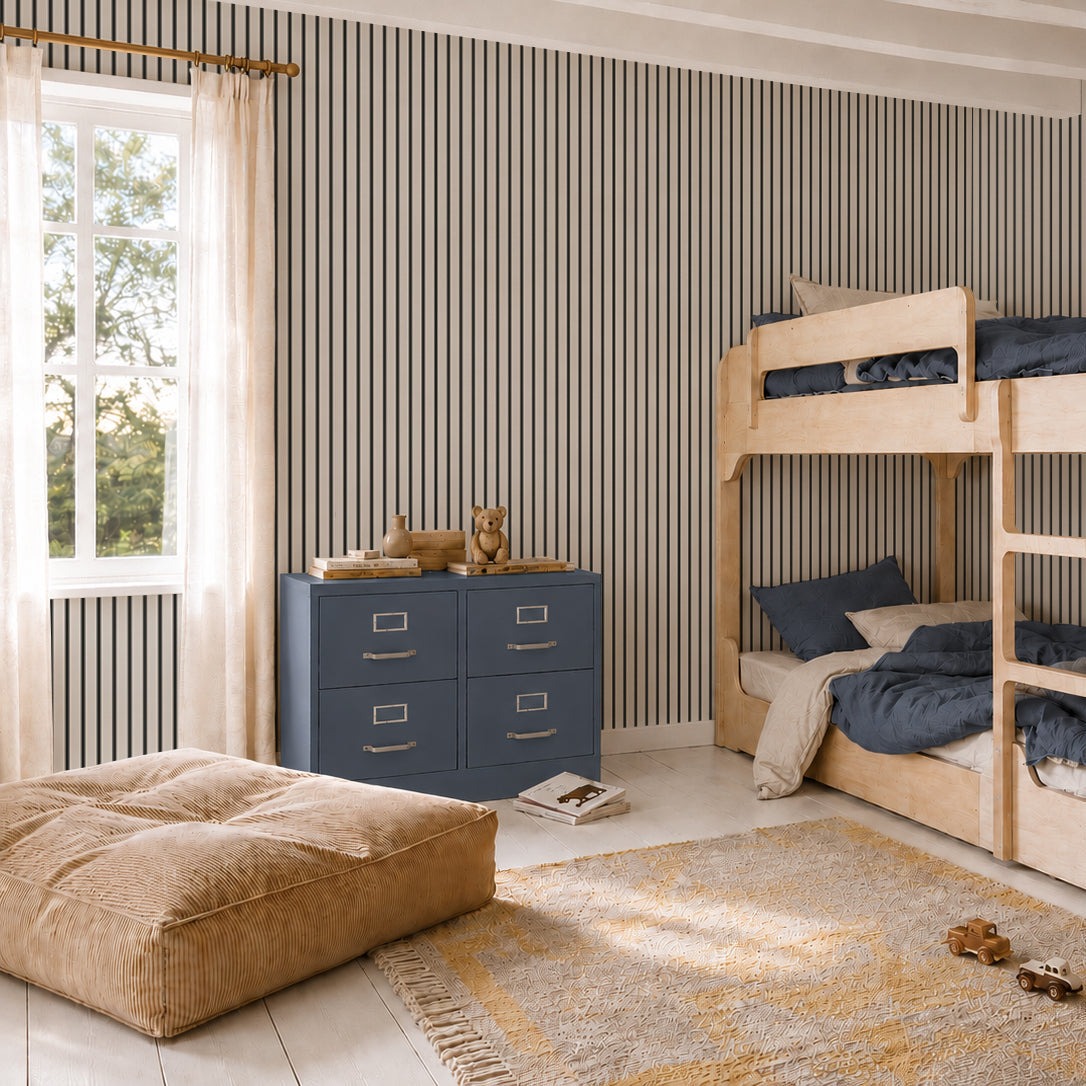 Children’s bedroom with wood bunk beds, a blue dresser with toys, a large tan pouf, and a yellow rug. The room features Pinstripe Wallpaper Charcoal for a playful touch and a window with sheer beige curtains letting in natural light—perfect for transitioning from nursery to kid’s space.