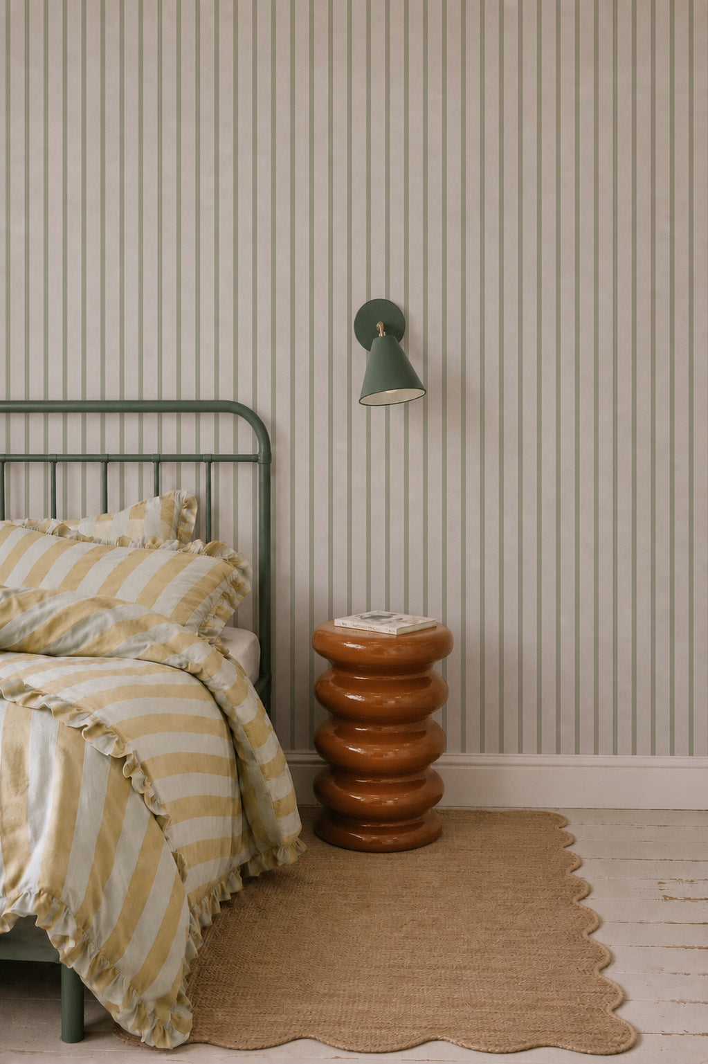 A cozy bedroom with a green metal bed frame, yellow and white striped bedding, a stacked round brown nightstand, a green wall sconce, Pinstripe Wallpaper Green, and a woven rug on a white wooden floor.