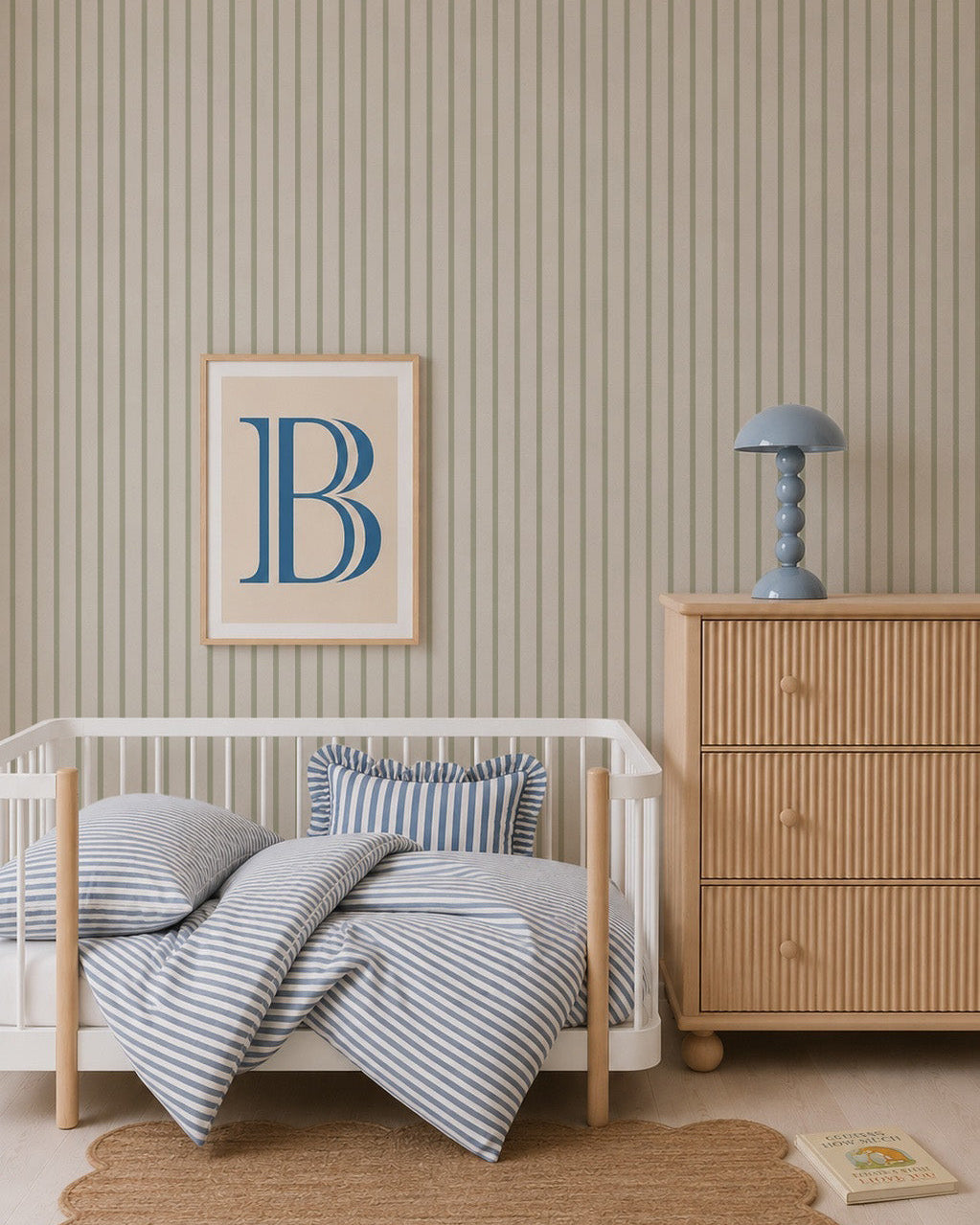A nursery with a white crib, blue and white striped bedding, Pinstripe Wallpaper Green, a chest of drawers with a blue lamp, a framed letter B on the wall, a round jute rug, and a children&