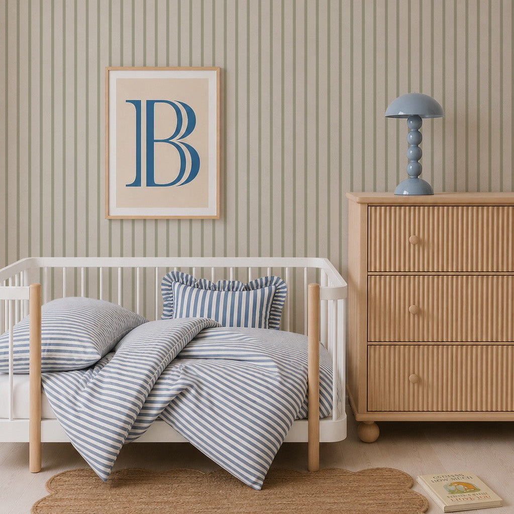 A nursery with a white crib, blue and white striped bedding, Pinstripe Wallpaper Green, a chest of drawers with a blue lamp, a framed letter B on the wall, a round jute rug, and a children&