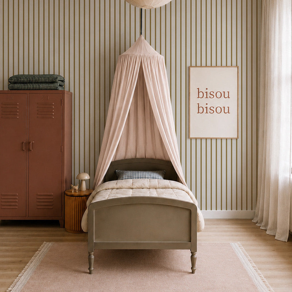 A cozy nursery features a single bed under a blush canopy, a large paper lantern, a pink wardrobe, framed "bisou bisou" art, and Pinstripe Wallpaper Moss adding warmth as sunlight filters through sheer curtains.