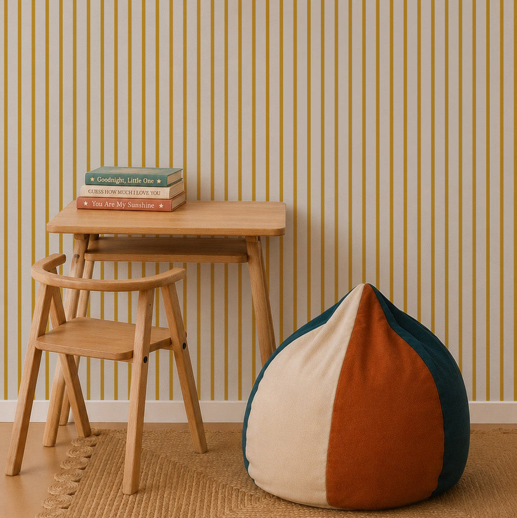 A minimalist room with a small wooden desk and chair, stacked books, a round beanbag in cream, orange, and green, and a hanging white lamp set against Pinstripe Wallpaper Mustard.