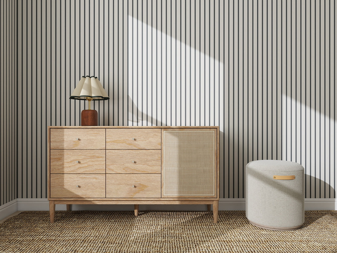 A wooden sideboard with drawers and a woven door stands against Pinstripe Wallpaper Denim, topped with a lamp. A textured ottoman sits to the right on a woven rug under bright sunlight.