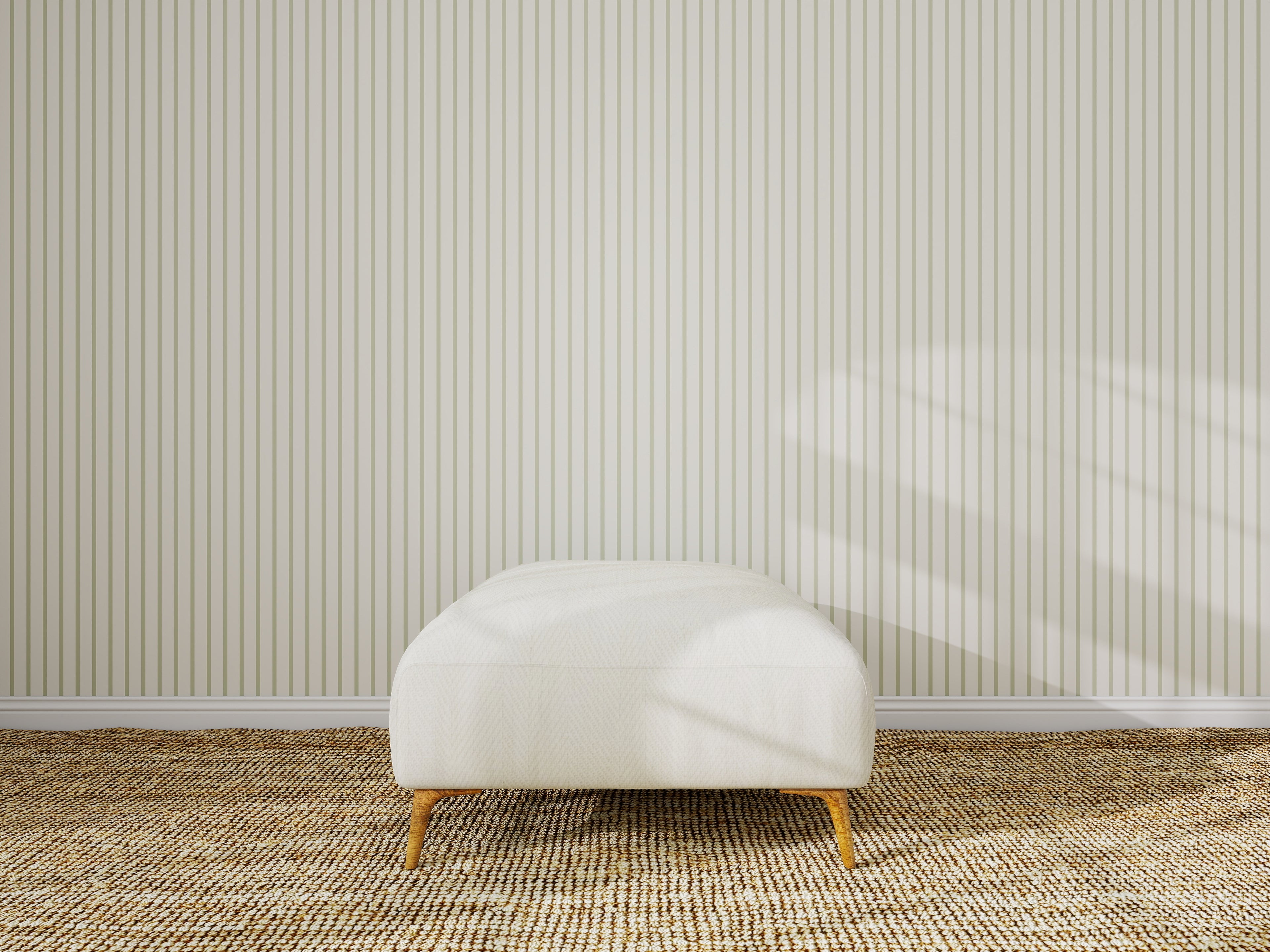 A minimalist room features a Pinstripe Green wallpaper sample on the wall, a textured tan carpet, and a single white ottoman with wooden legs in the center. Sunlight casts gentle shadows on the wall.