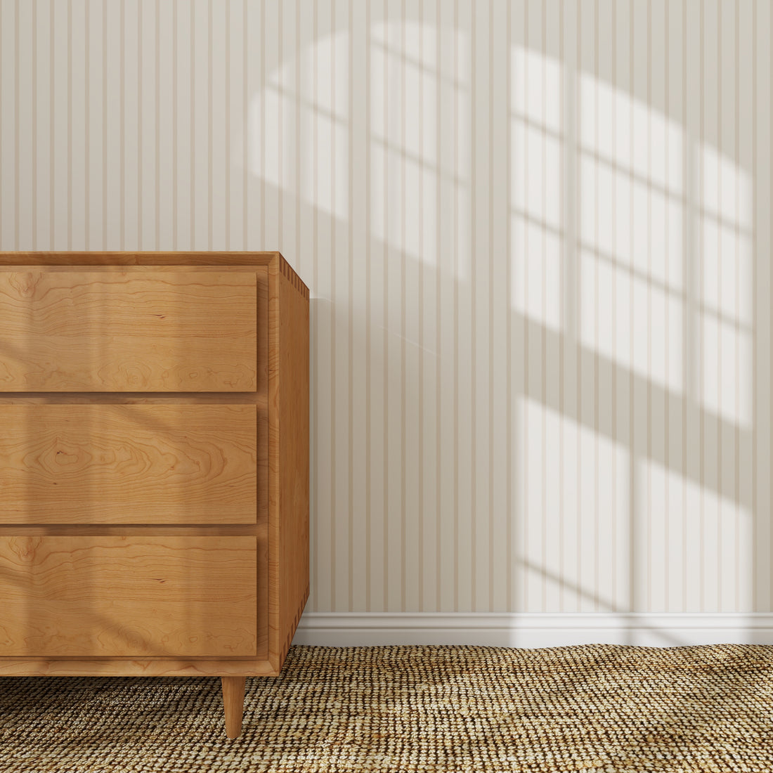 A wooden dresser with three drawers sits on a woven rug beside the Pinstripe Wallpaper Sample in Soft Beige. Sunlight casts a window-shaped shadow, emphasizing the wallpaper&