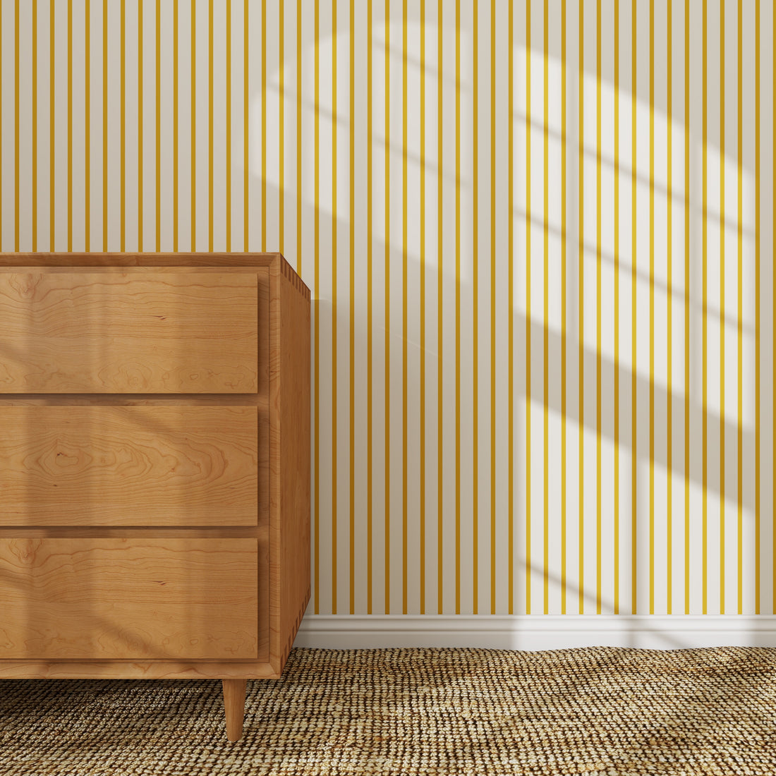 A wooden dresser stands on a woven rug beside a wall decorated with Pinstripe Wallpaper Mustard. Sunlight streams through the window, casting gentle shadows and adding a cozy nursery touch.
