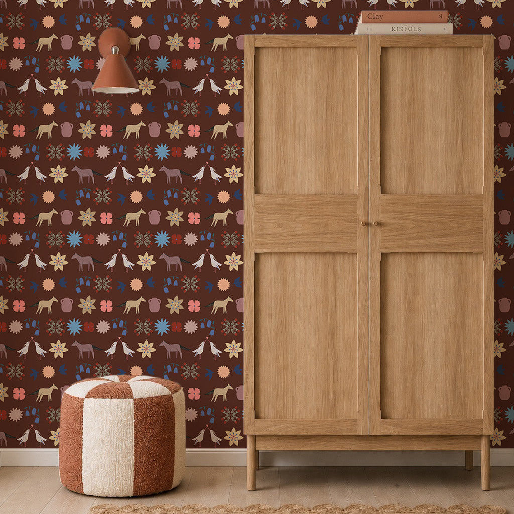 A wooden wardrobe stands on a woven rug beside a round, red and white pouf. The dark brown wall is decorated with Playful Prairie Cocoa Wallpaper, featuring whimsical animal and floral patterns. A small wall lamp is mounted above the pouf.