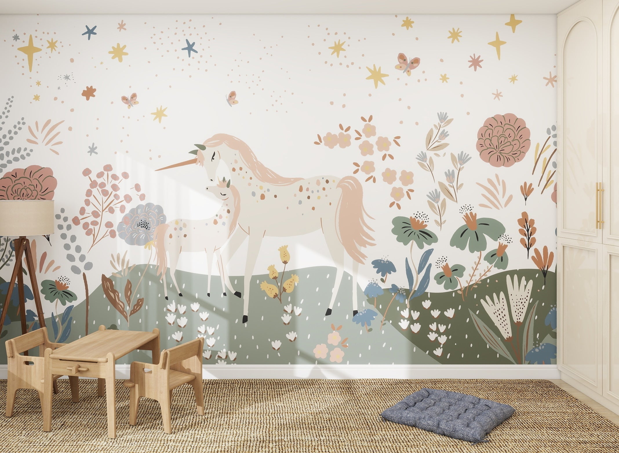 A children’s room featuring the Unicorn Meadow Wallpaper Mural, showing two unicorns among colorful flowers, leaves, and stars. The space includes a small wooden table with chairs, a blue floor cushion, a lamp, and built-in white cabinets.
