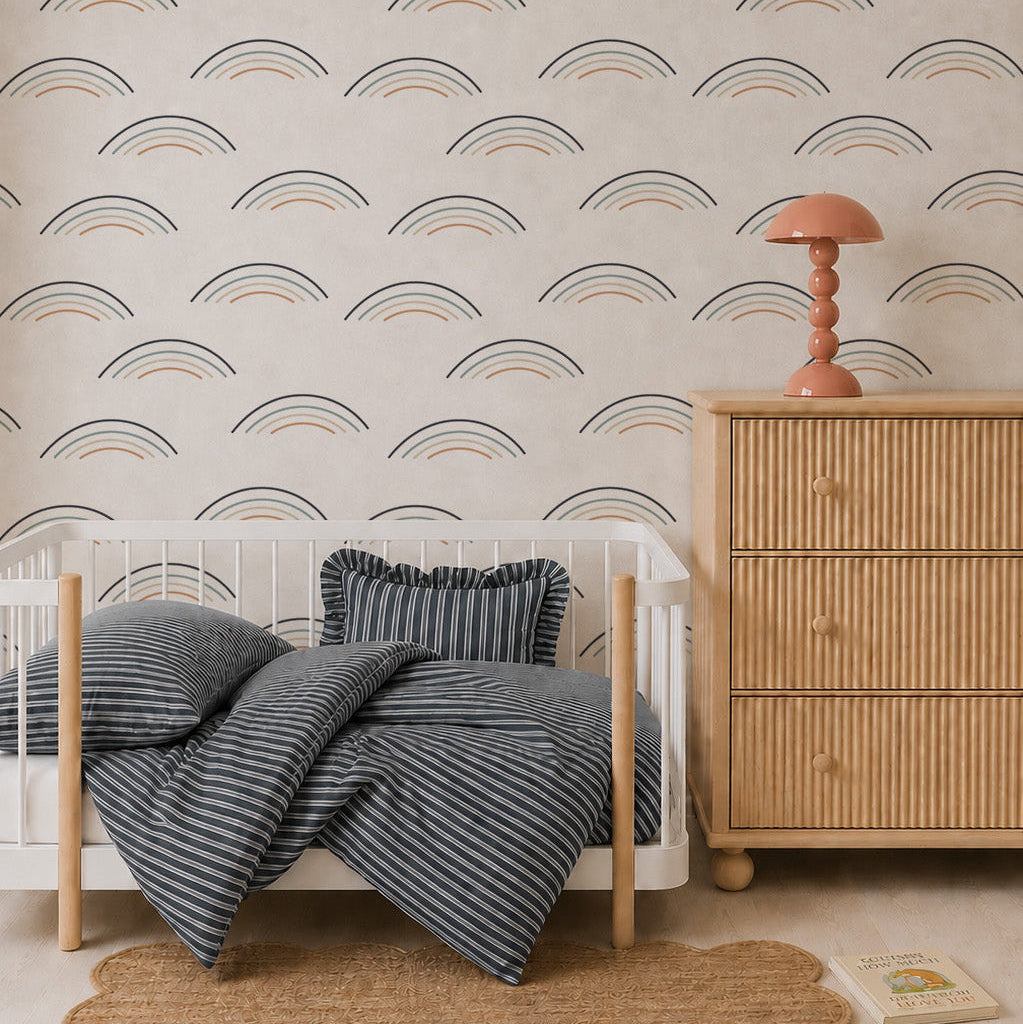 A cozy nursery with a white crib, blue striped bedding and pillow, rattan dresser with a coral lamp, scalloped rug, Astrid Unicorn Wallpaper, and a children’s book on the floor.