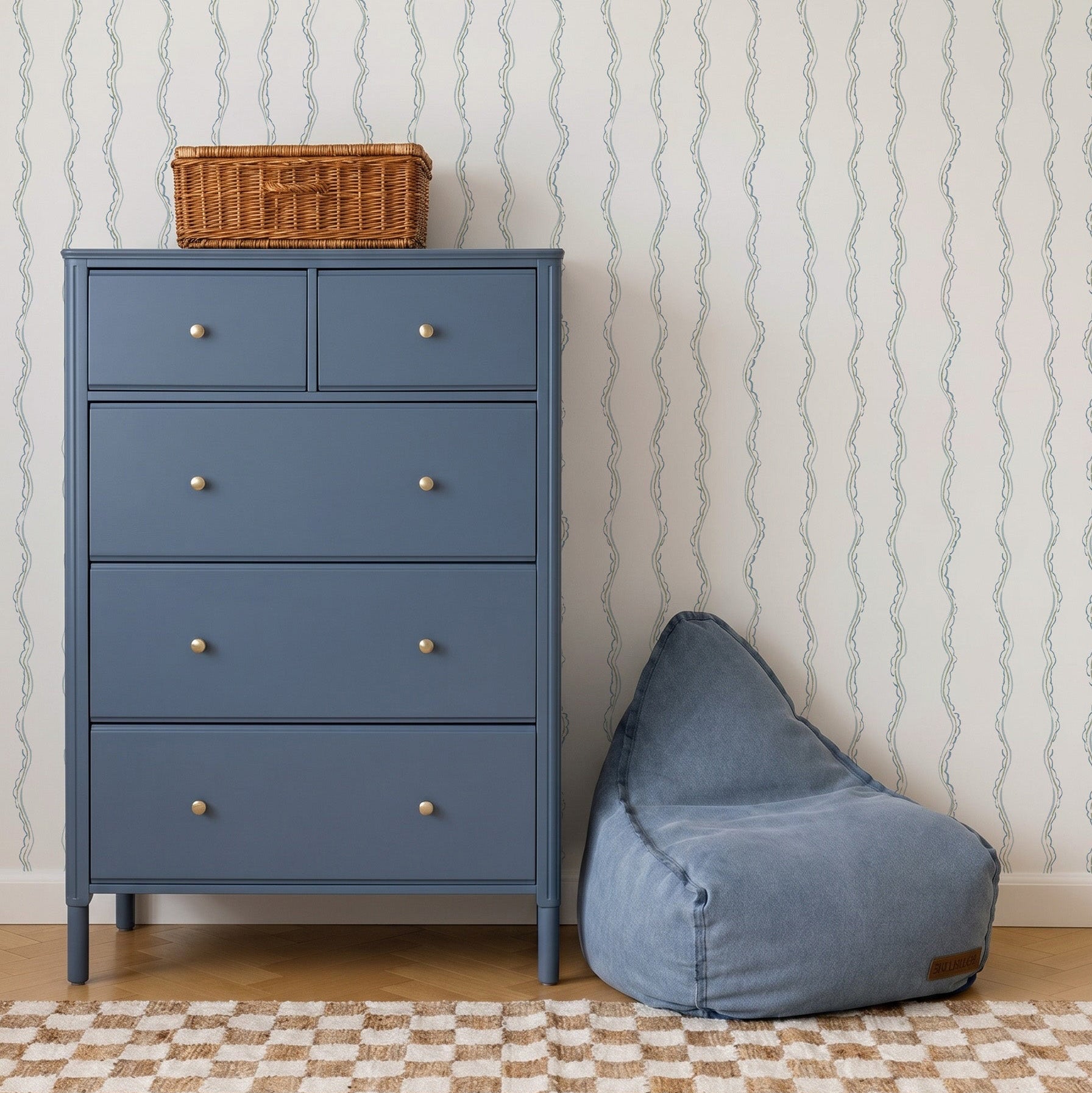 A blue dresser with four drawers and a wicker basket on top stands next to a blue bean bag chair. The background features Ribbon Stripe Wallpaper in Blue and a beige checkered rug, creating calming nursery decor.