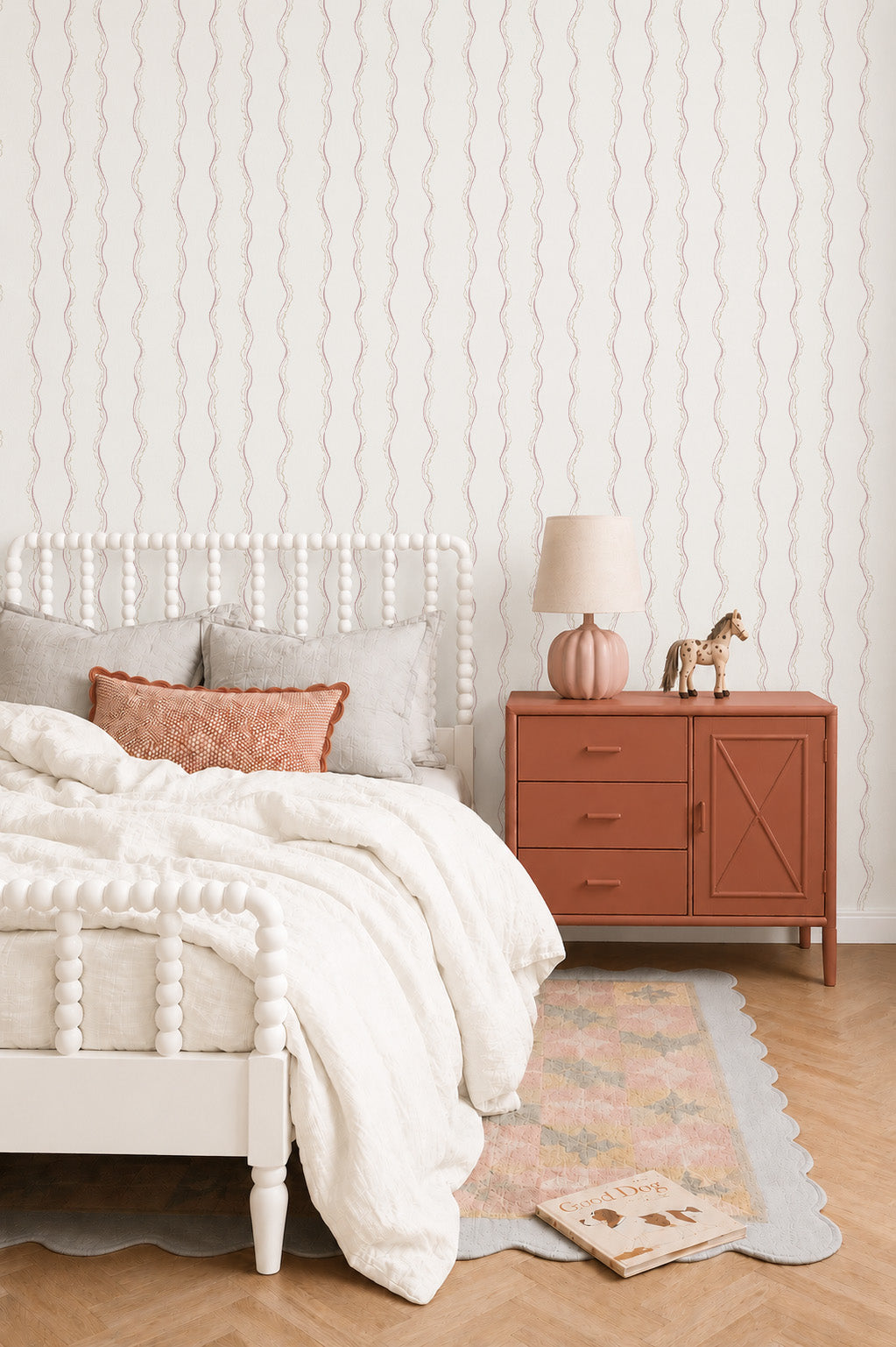 A cozy bedroom features a white spindle bed with neutral bedding, a peach accent pillow, Ribbon Stripe Wallpaper in Rose, a rust nightstand with lamp and toy giraffe, plus a pastel rug on light wood floors.