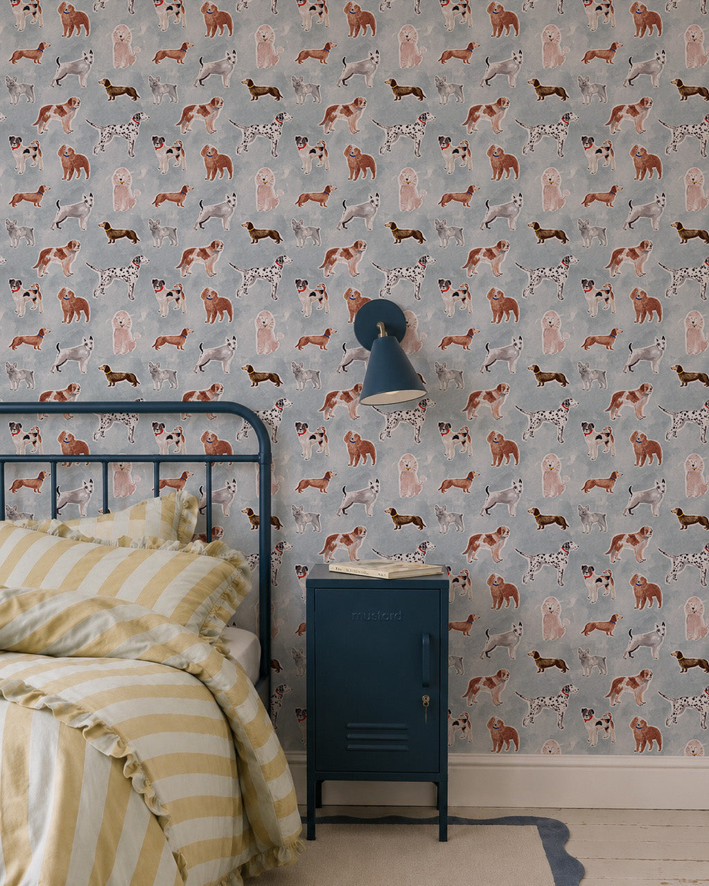 A cozy bedroom features a metal-framed bed, yellow and white striped bedding, a blue nightstand, a wall lamp, and cheerful Rigby Dog Print Wallpaper in blue on a gray background.
