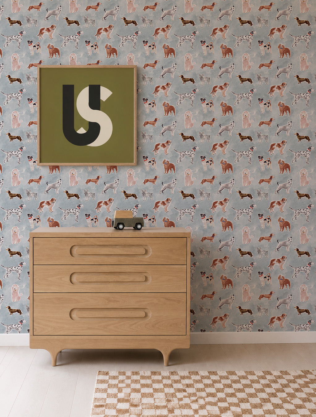 Rigby Dog Print Wallpaper, Blue featuring a repeating pattern of various dog breeds on a blue background, displayed behind a wooden dresser and a framed graphic print.