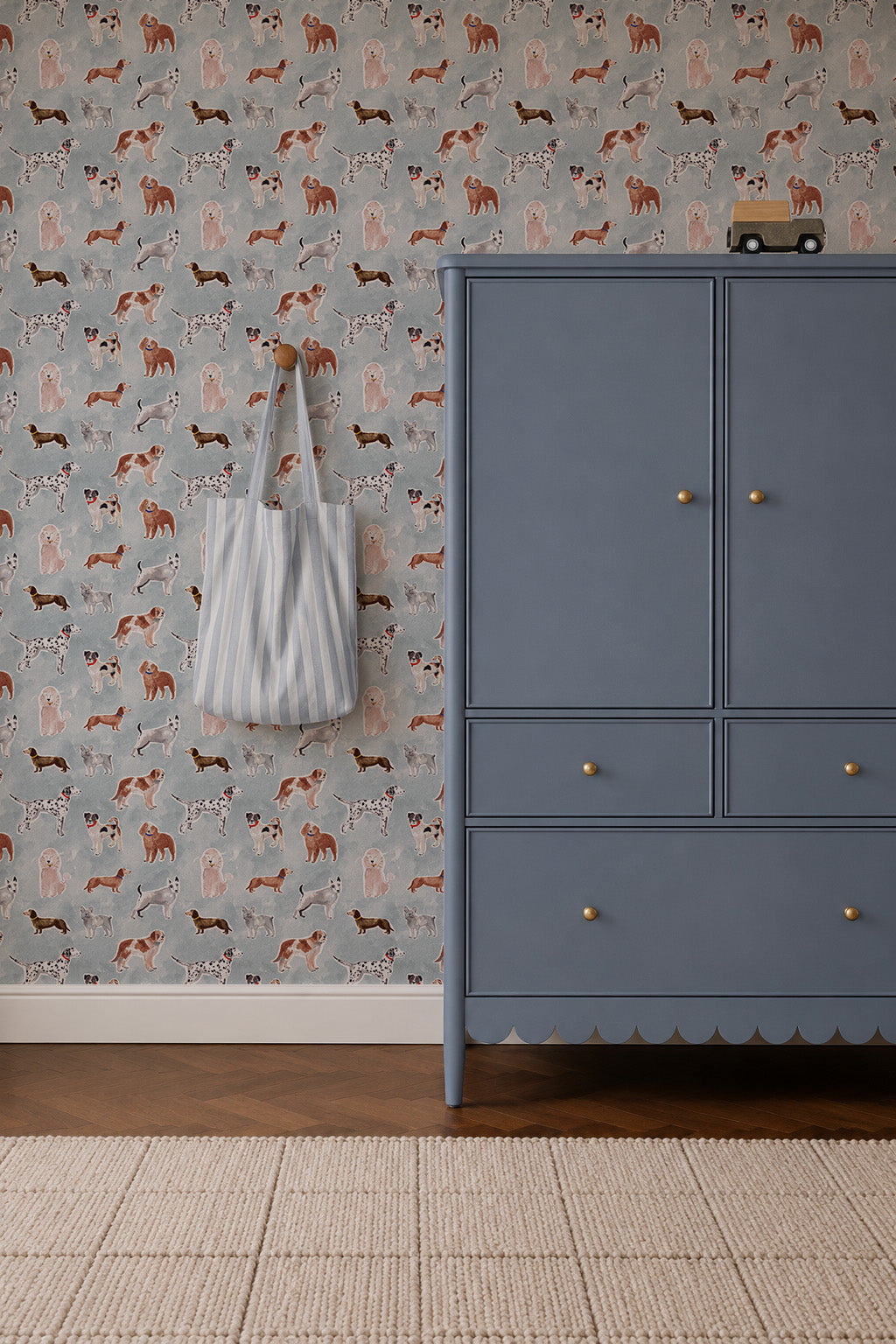 Rigby Dog Print Wallpaper, Blue featuring a repeating pattern of various dog breeds on a blue background, next to a blue armoire and a striped tote bag hanging on the wall.