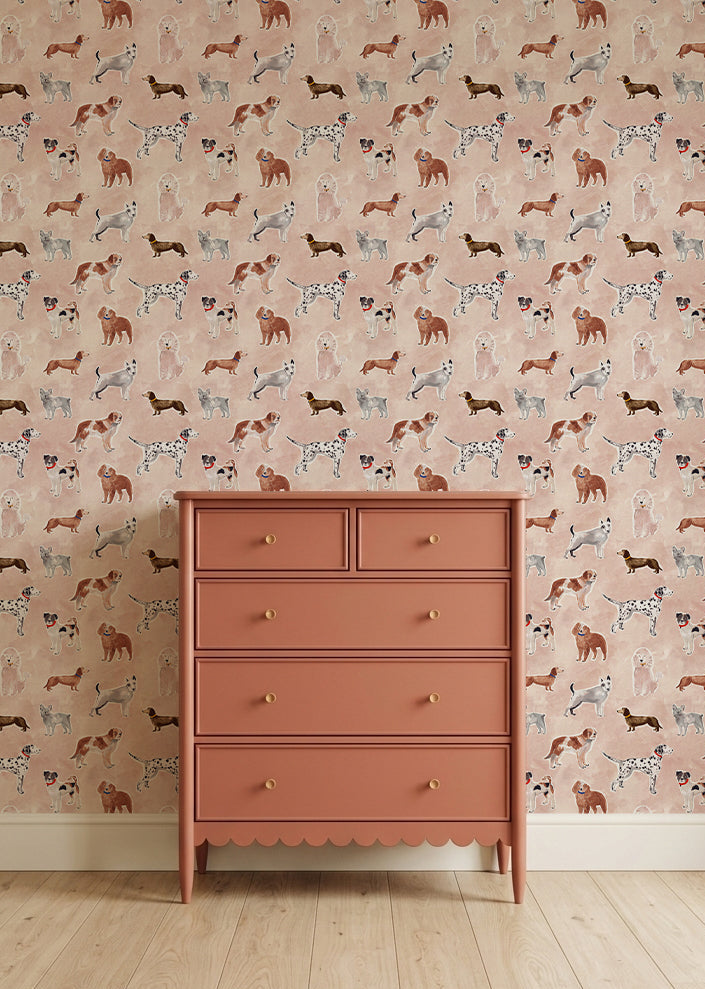 A coral-colored five-drawer dresser stands on a light wooden floor against a wall covered with Rigby Dog Print Wallpaper in Rose, featuring whimsical pink, brown, black, and white dog motifs.