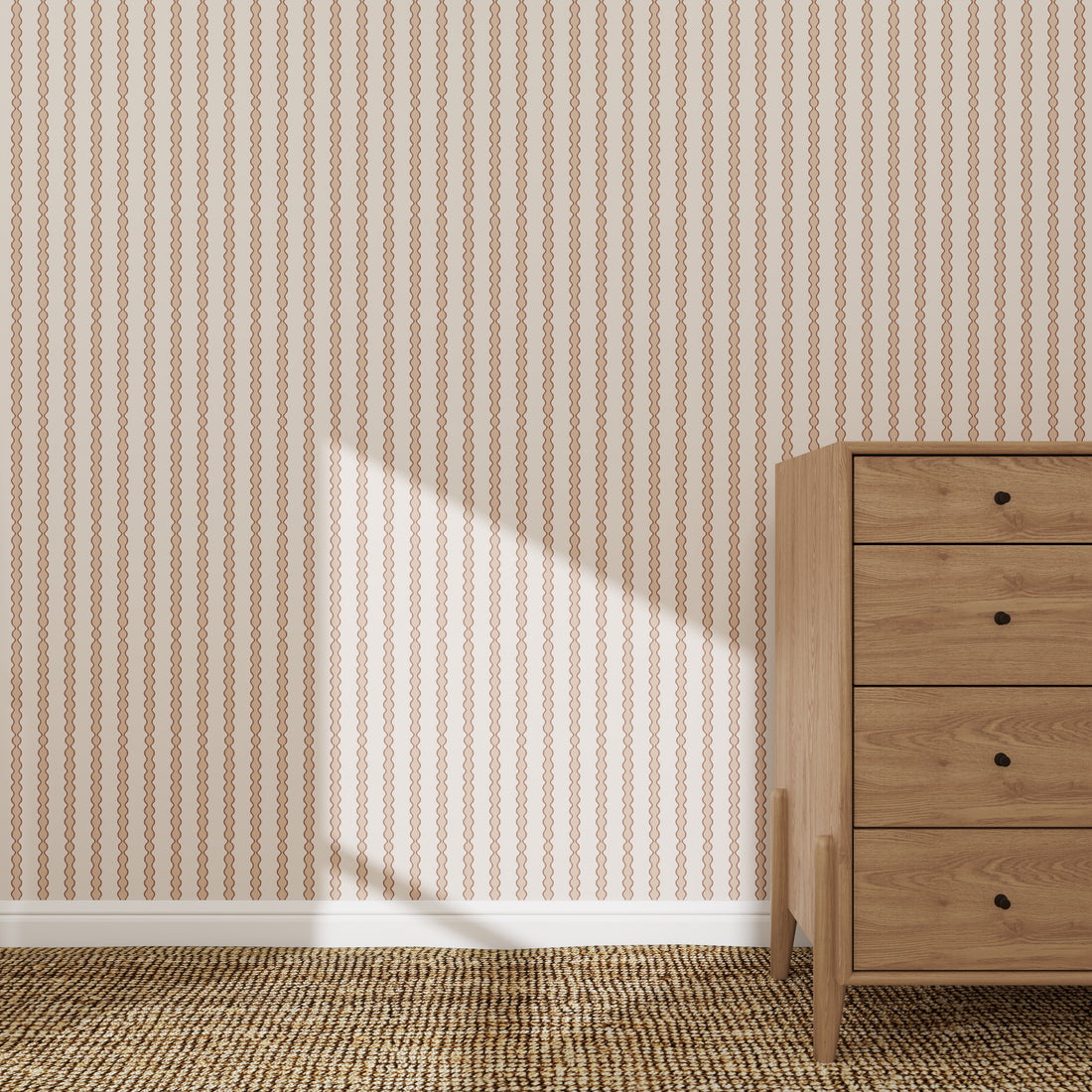 A wooden dresser with five drawers sits on a woven carpet beside a wall adorned with Little Abacus Stripe Wallpaper Sample in Rose. Sunlight casts a triangular shadow, highlighting the elegant pattern and quality of the wallpaper.