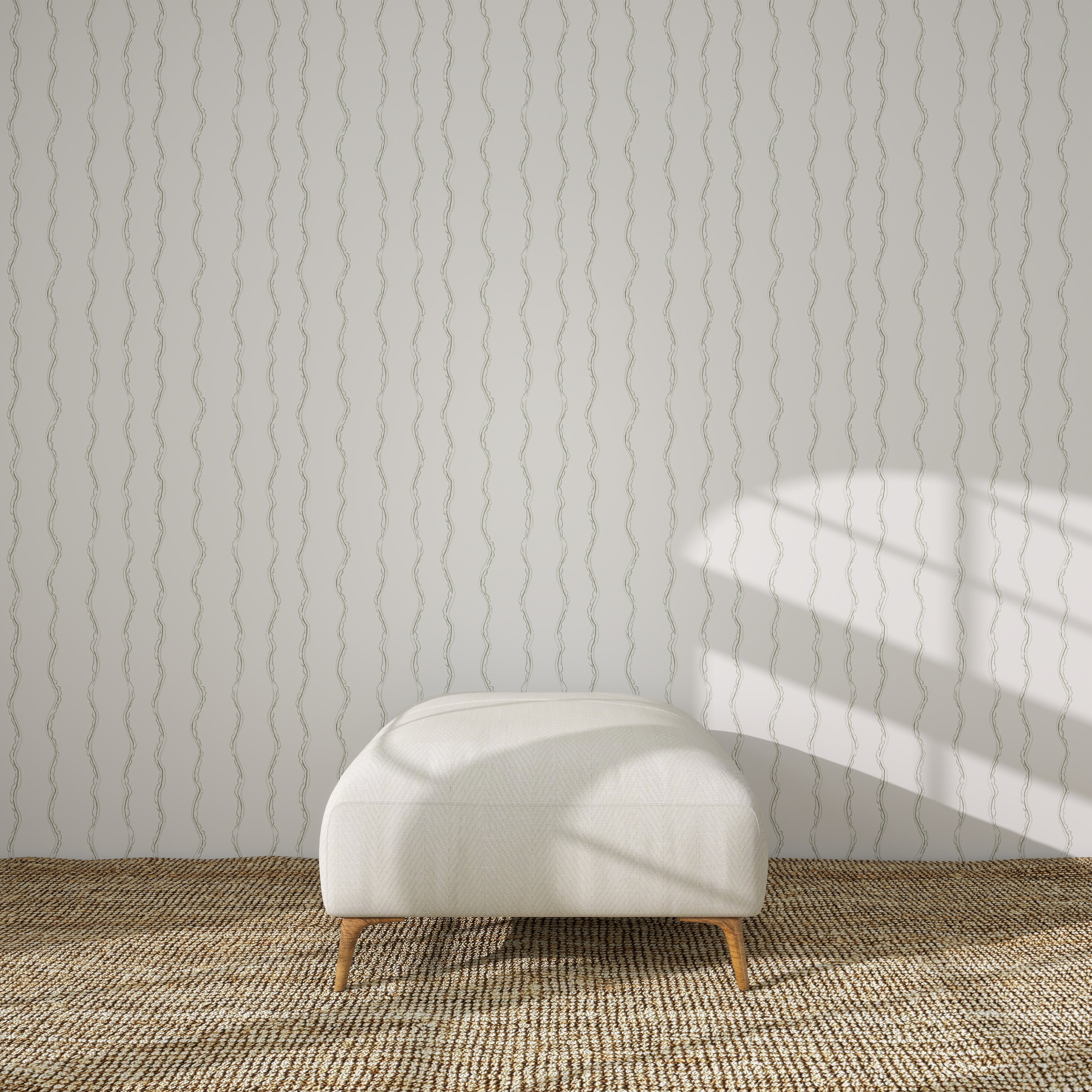 A modern white ottoman with wooden legs sits on a woven beige rug in front of a light gray wall decorated with Ribbon Stripe Wallpaper in Sage. Sunlight and shadows fall across the wall from the right.