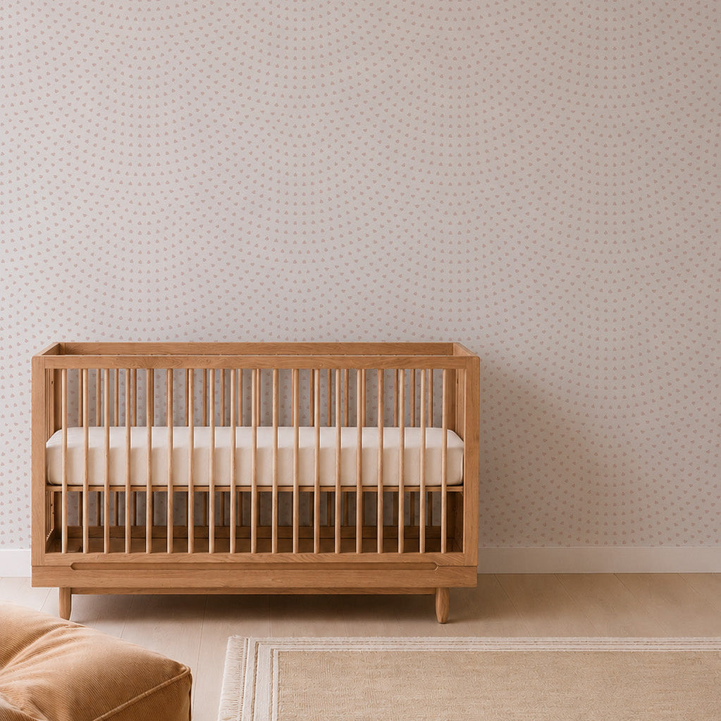 A wooden baby crib with white bedding stands against a light wall adorned with the Scallop Shell Mural in Pink, adding a subtle coastal touch to the nursery. There is a beige rug on the light wood floor in front of the crib, enhancing the serene and minimalist décor of the space.
