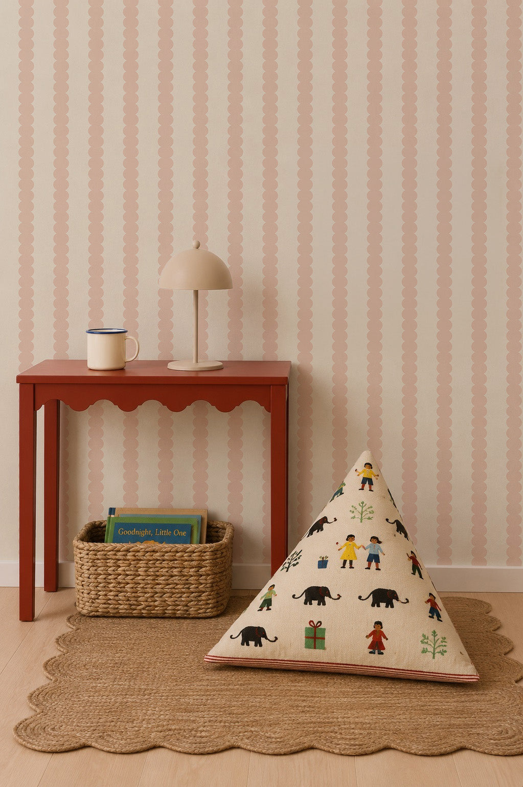 A red table with a lamp and mug sits against blush Scallop Stripe Wallpaper. Below, a woven basket holds two books, and a triangular cushion with colorful animals rests on a scalloped rug.