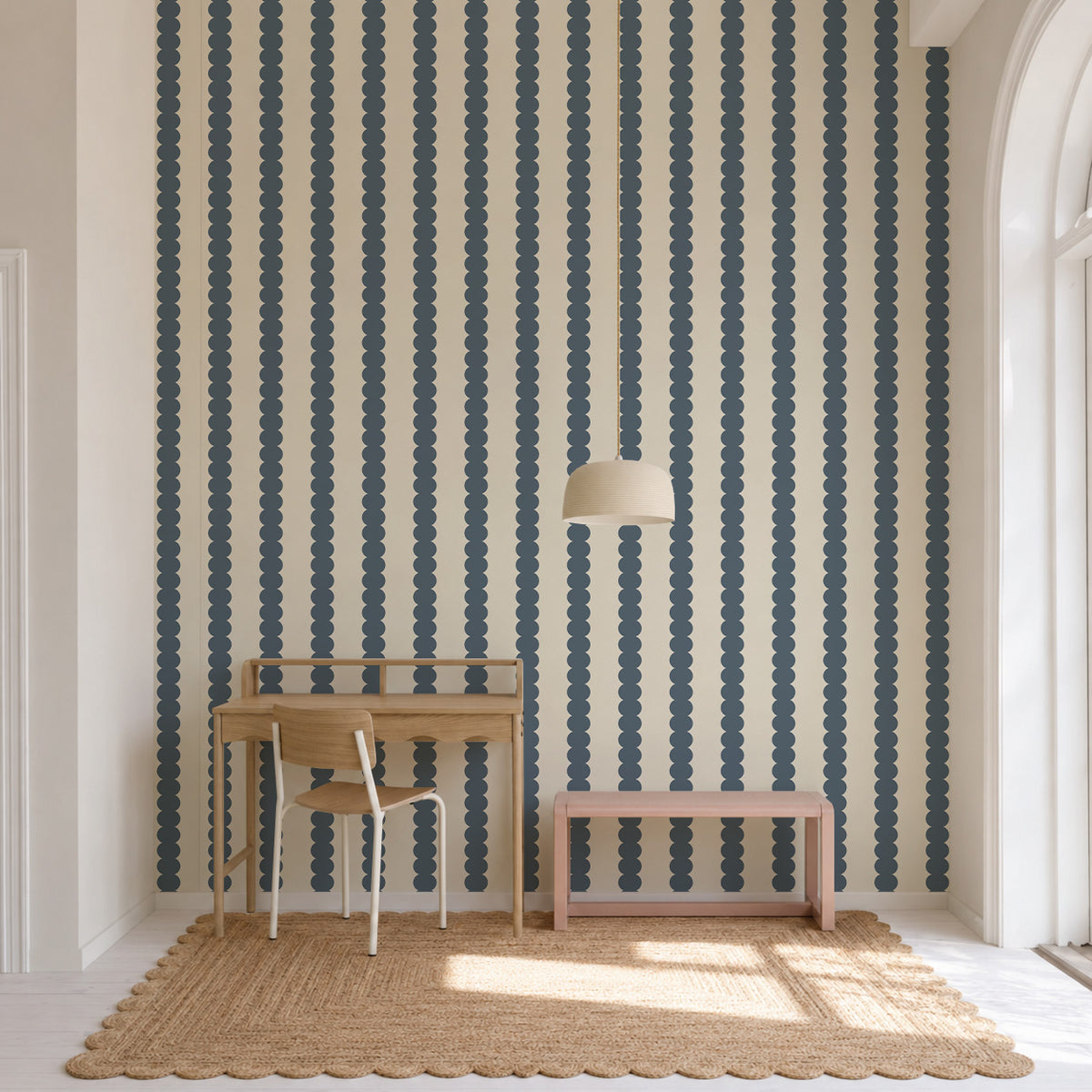 A wooden desk, chair, and pink bench set against Scallop Stripe Wallpaper, Deep Blue, featuring vertical rows of scalloped circles on a light cream background.