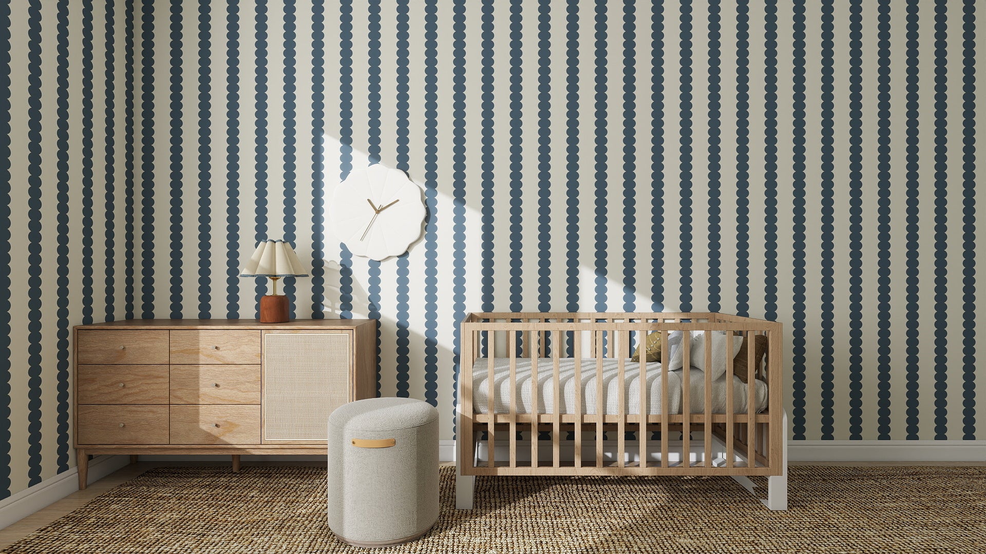 A modern nursery with a wooden crib, chic decor, and a small ottoman features Scallop Stripe Wallpaper Deep Blue in a bold dotted pattern. A woven rug brings warmth to the cozy blue-and-white palette.