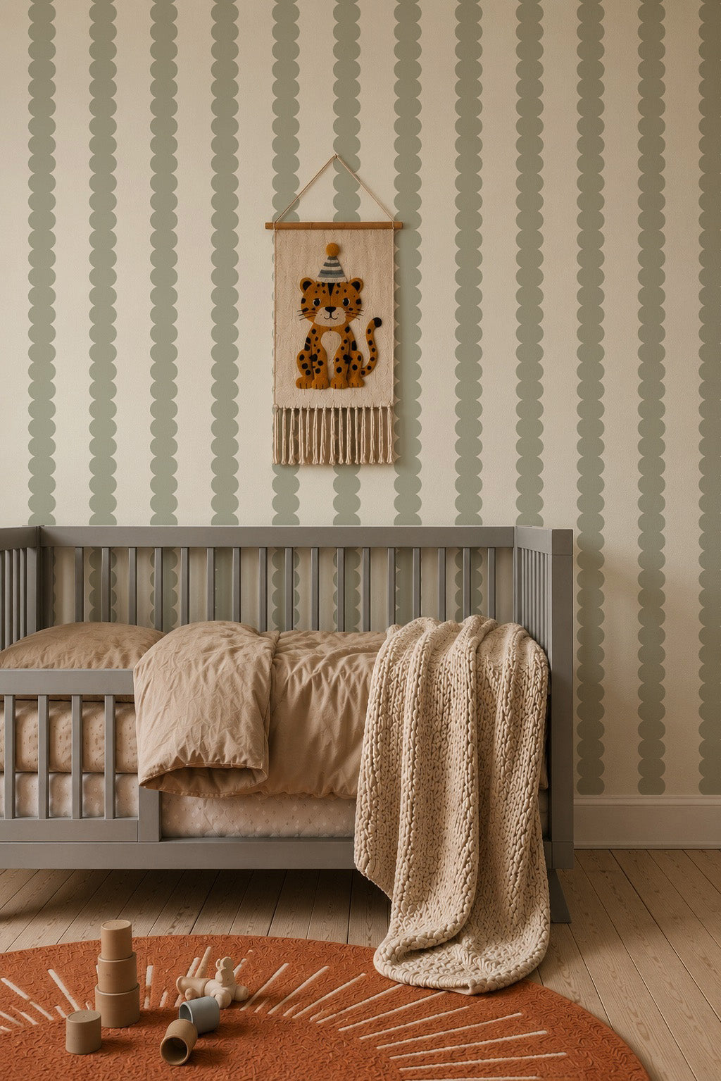 A cozy nursery with a gray crib, tan bedding, a chunky knit blanket, a rust rug, and wooden toys. The walls feature elegant Scallop Stripe Wallpaper in green, which beautifully frames the decorative hanging of a spotted animal in a hat.