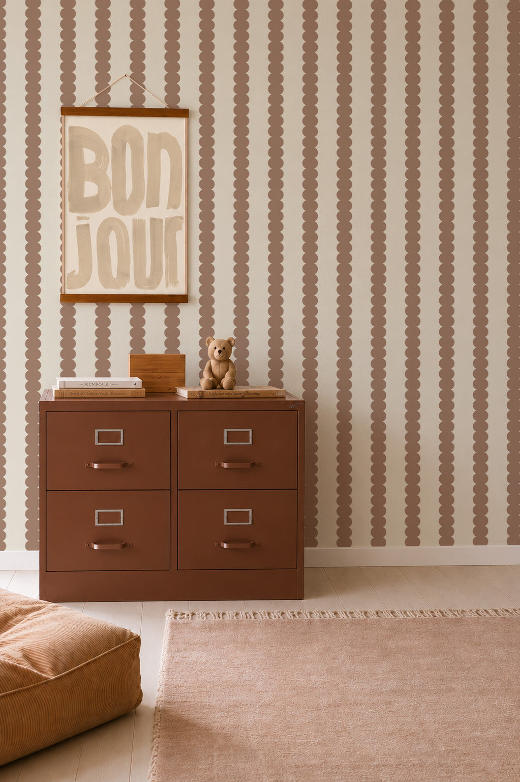 A brown cabinet with four drawers stands against a patterned wall featuring the Scallop Stripe Wallpaper in Latte, with a BONJOUR sign above it. A small bear and books sit on top. Nearby are a tan pouf and a fringed rug on a light floor.