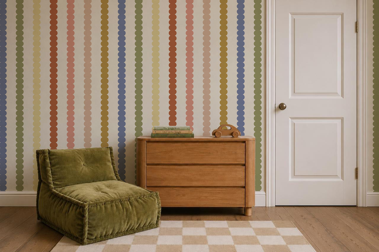 A cozy room with a green chair, a wooden dresser holding books and a toy, a checkered rug, and a door. The wall features the Scallop Stripe Wallpaper in Multi, showcasing vertical wavy stripes in muted colors for an inviting touch.
