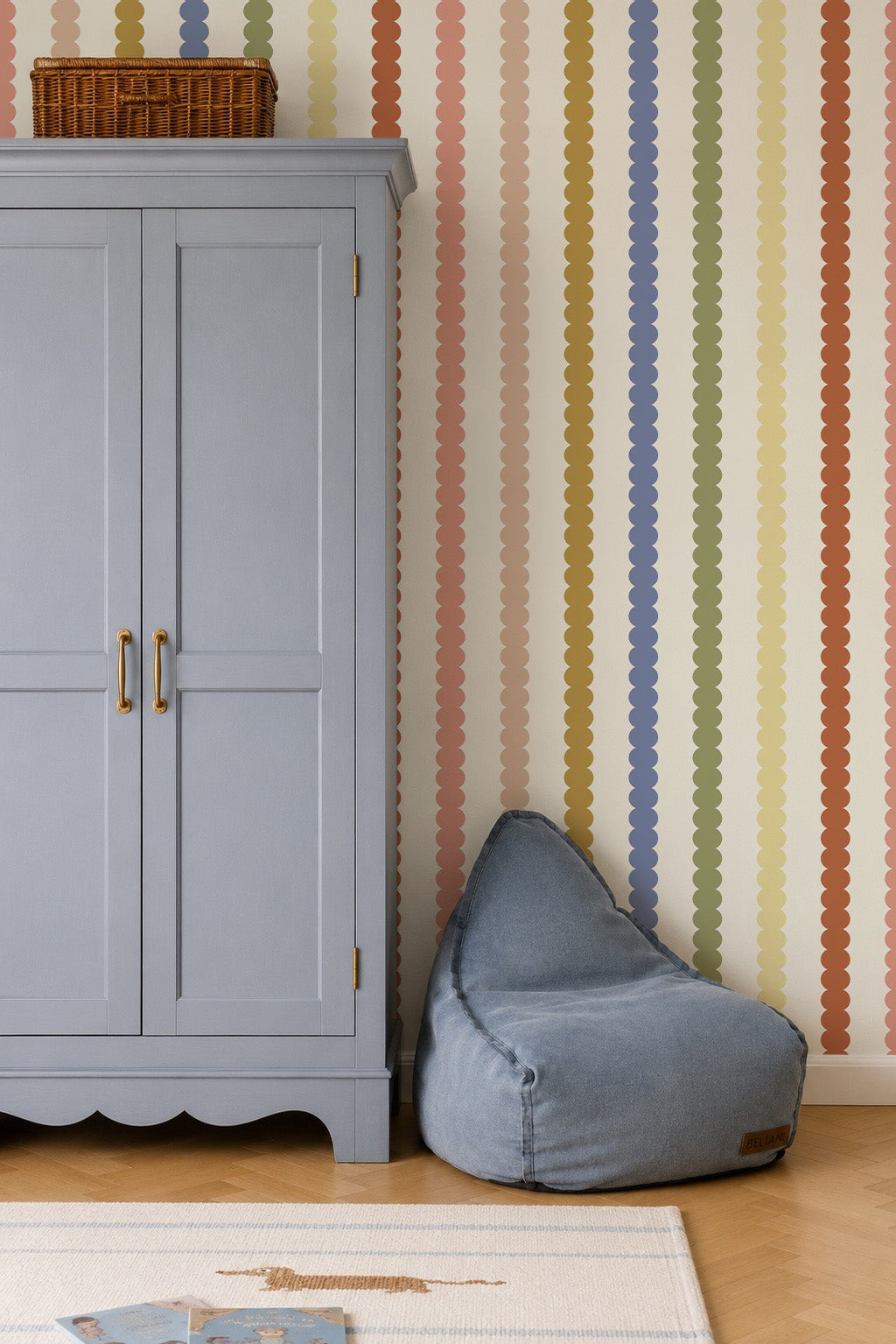 A light blue wardrobe stands next to a blue bean bag chair. The wall behind them features vertical, wavy stripes in various colors, created by Scallop Stripe Wallpaper, Multi. A wicker basket sits atop the wardrobe, and part of a rug is visible on the wooden floor.