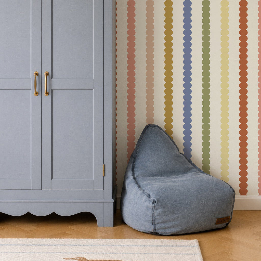 A light blue wardrobe stands next to a blue bean bag chair. The wall behind them features vertical, wavy stripes in various colors, created by Scallop Stripe Wallpaper, Multi. A wicker basket sits atop the wardrobe, and part of a rug is visible on the wooden floor.