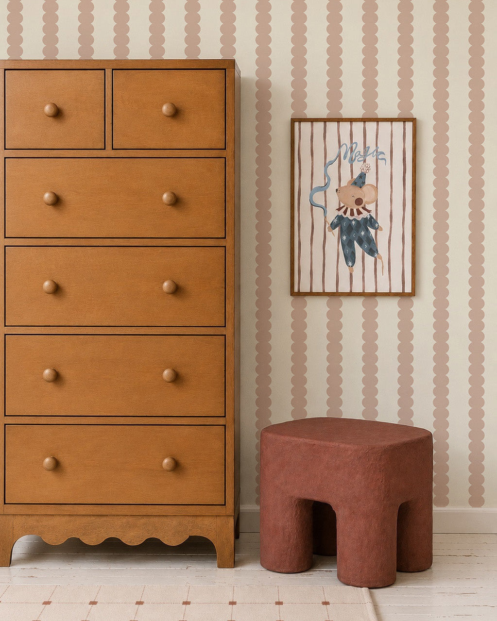A wooden dresser with round knobs stands by a reddish-brown stool and framed art on a wall covered in Scallop Stripe Wallpaper, Pink. The light grid-patterned floor completes this playful kids’ room setup.