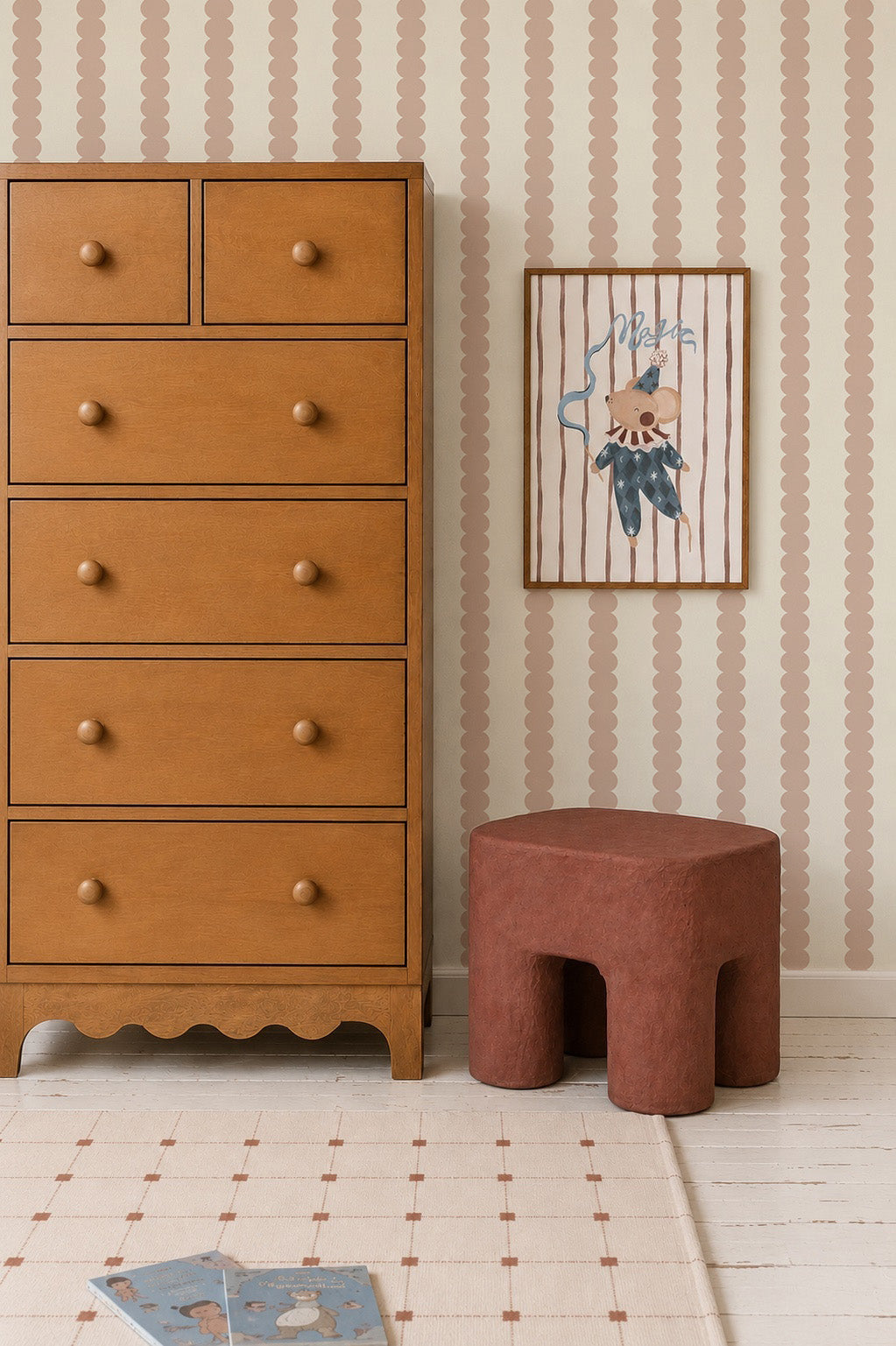 A wooden dresser stands next to a brown stool under a framed illustration of a mouse on Scallop Stripe Wallpaper, Pink. An open children’s book lies on a cream rug with a grid pattern, adding charm to this cozy kids room.
