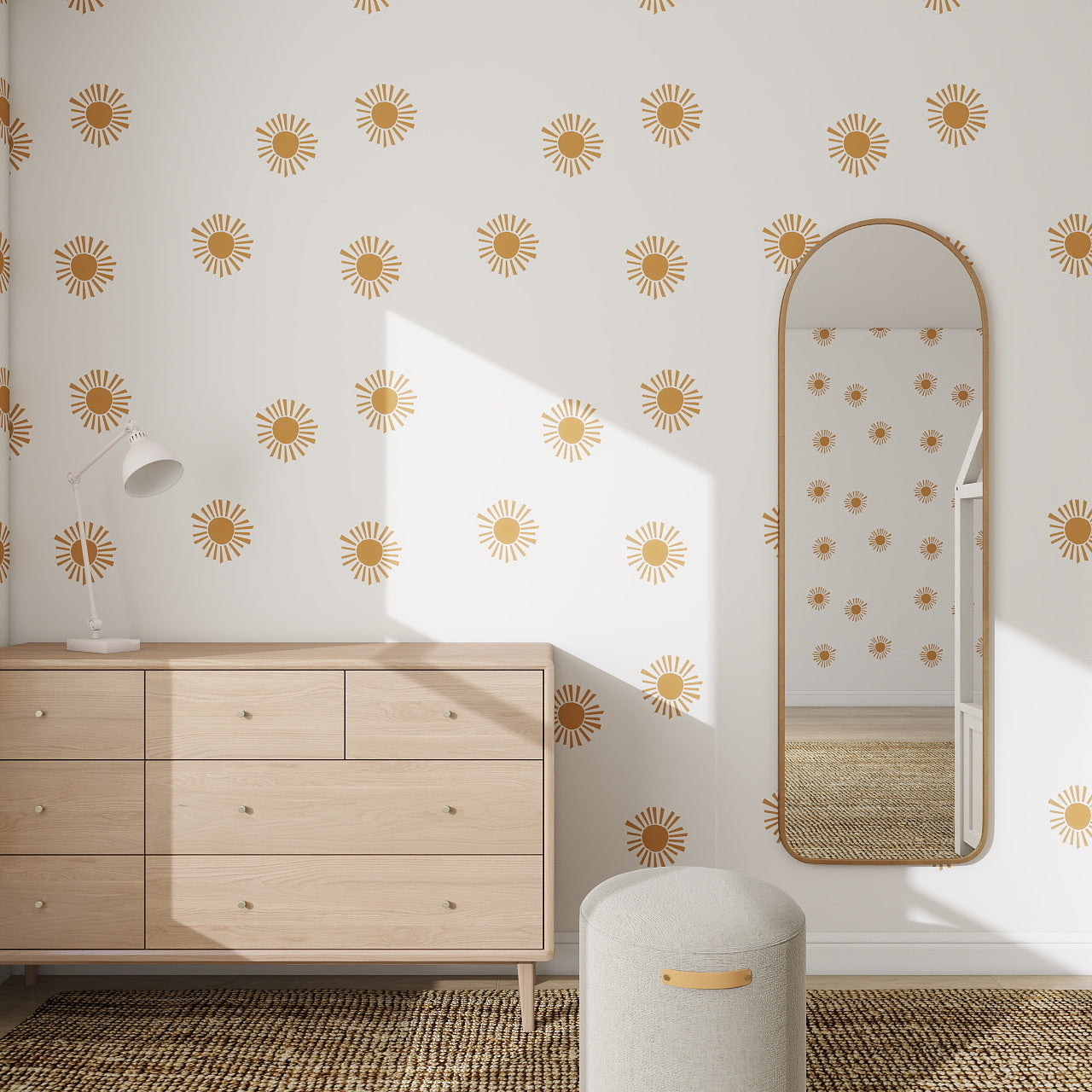 A minimal bedroom with white walls features the Shining Sun Wallpaper Repeat Pattern in a gold sunburst design, a wooden dresser, tall arched mirror, and light gray ottoman on a woven rug. Sunlight streams in, highlighting the unique decor.