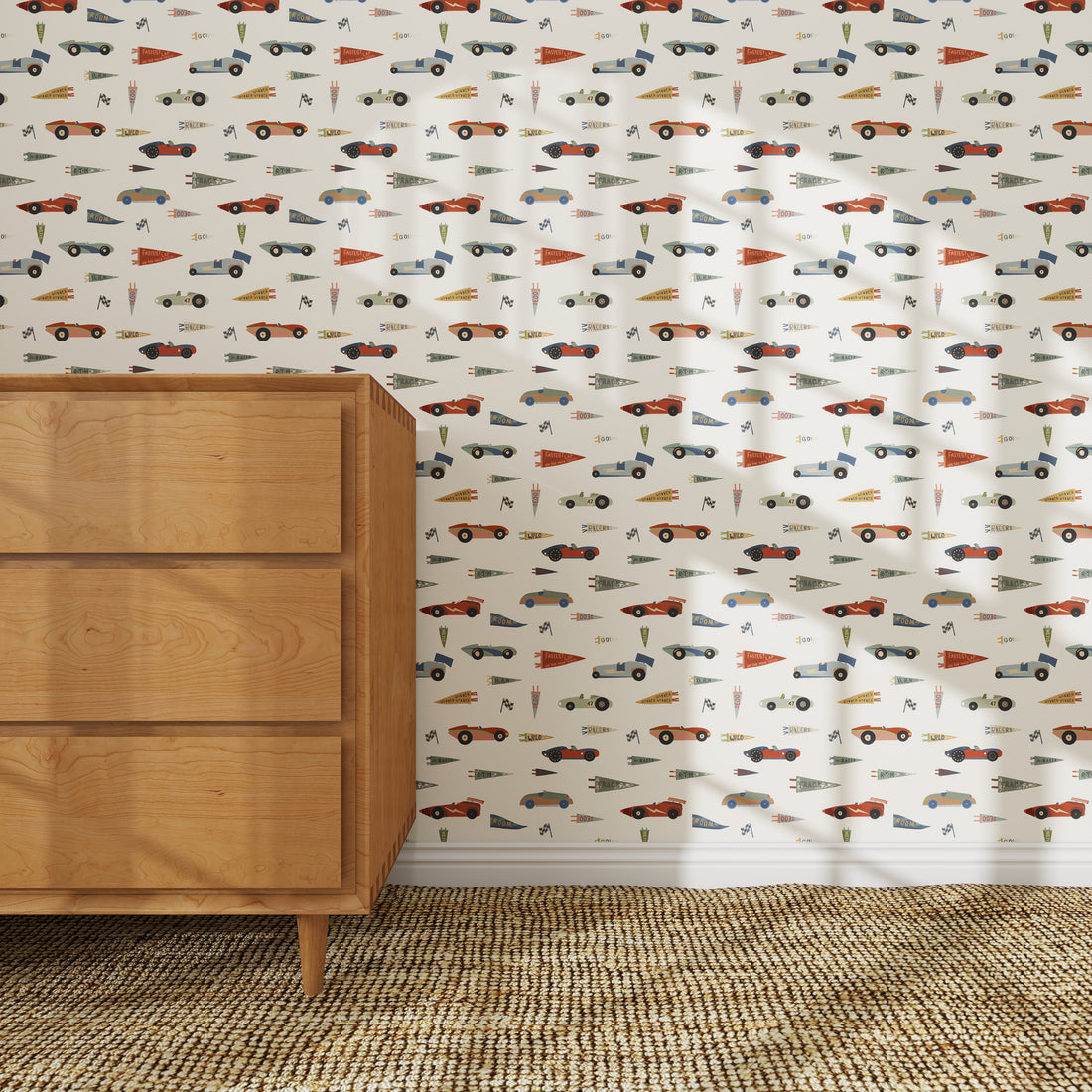 A light-filled room with a woven rug, a wooden mid-century dresser, and nursery wall decor featuring colorful vintage cars and pennants on Speedway Dash Wallpaper with a white background.