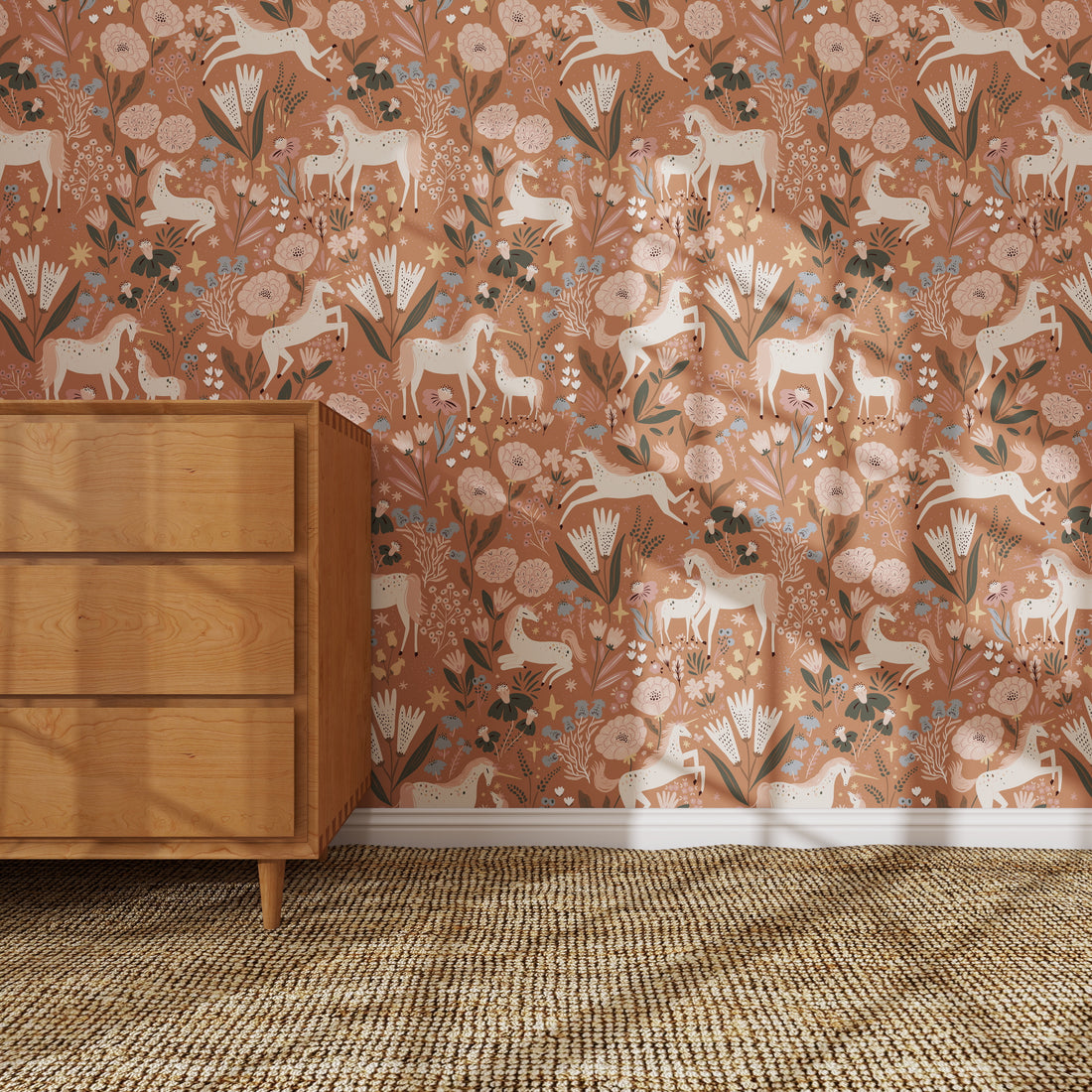 A wooden dresser stands on a woven rug by a wall covered in Unicorn Meadow Wallpaper Toffee Rose, featuring whimsical white deer, birds, flowers, and leaves in peach, pink, and green—perfect for charming girly room decor.