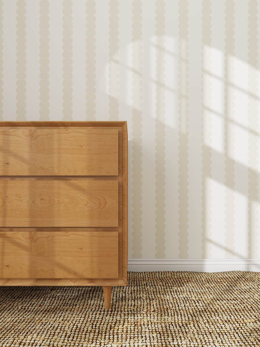 A wooden dresser with three drawers stands on a woven rug beside a wall adorned with Scallop Stripe Wallpaper Cloud, as sunlight streams in and casts window shadows across the wall and floor.
