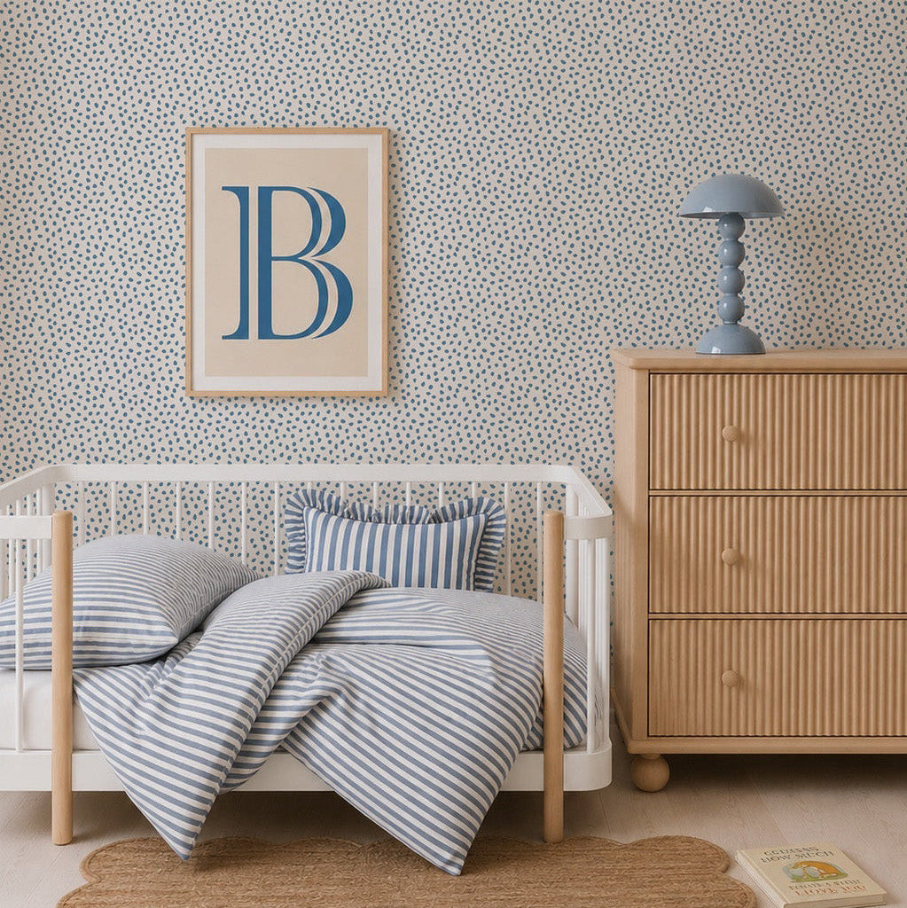A cozy nursery features a white crib, blue striped bedding, wooden dresser, blue lamp, framed art with a large blue B, a round woven rug, and Sprinkle Wallpaper in Blue. A book lies on the floor to complete the inviting scene.