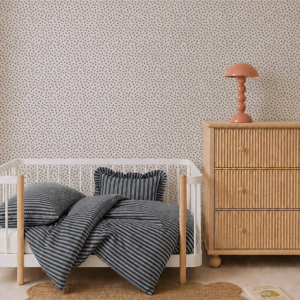 A minimal nursery with a white crib, striped bedding, a wooden dresser topped with a pink lamp, a scalloped rug, Sprinkle Wallpaper in Grey featuring a repeating pattern, and a book on the floor showcases thoughtful interior design.